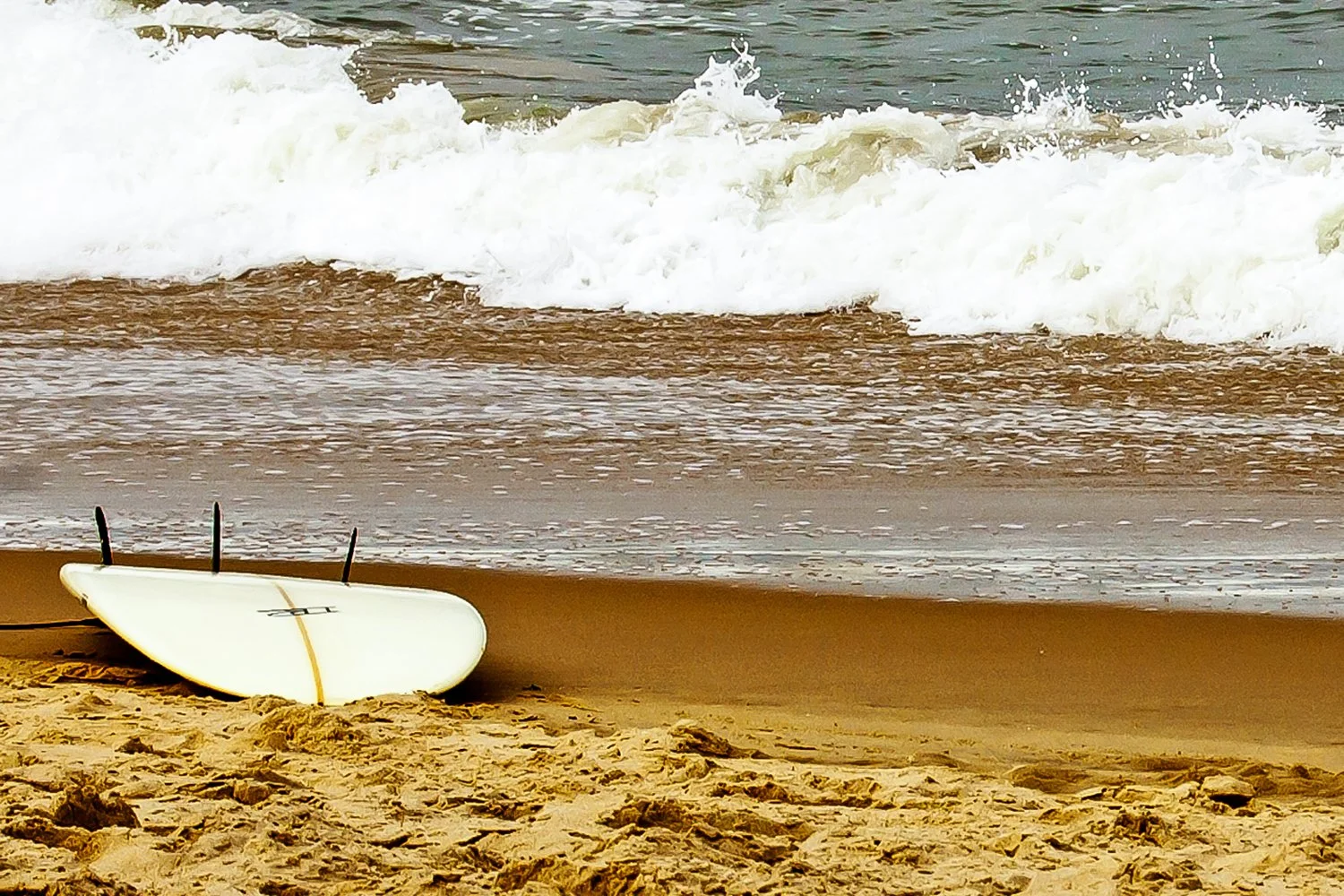 A white surfboard with three black fins lying on the sandy beach near the water's edge, with small waves crashing on the shore in the background.