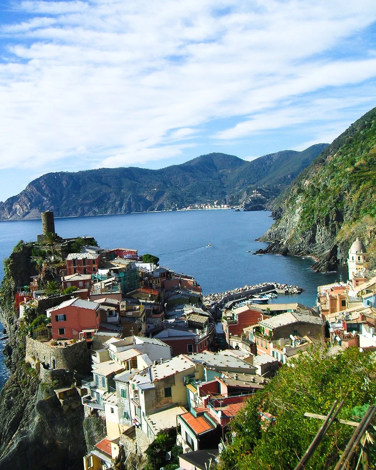 Colorful houses built on a rocky hillside overlooking the sea, with mountains in the background and cloudy blue sky above.