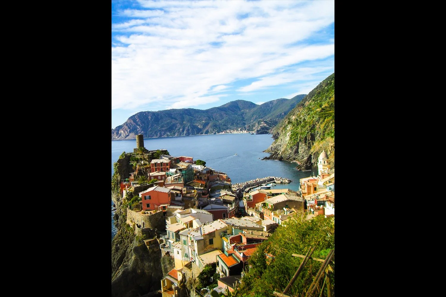 Colorful coastal village with houses on a hillside overlooking a bay, with mountains in the background and a partly cloudy sky.