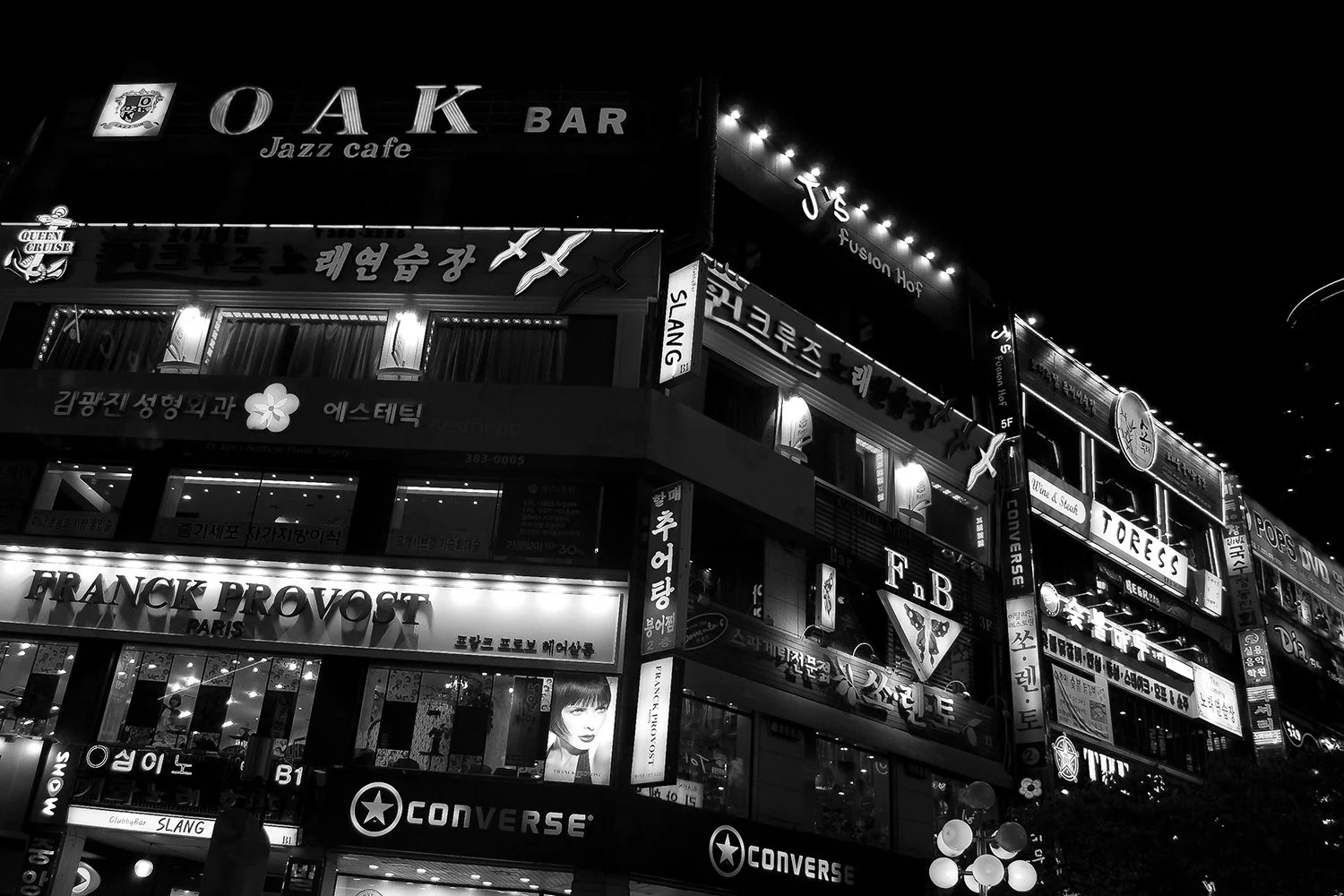 Night view of a multi-story building with brightly lit signs for cafes, bars, and shops in both Korean and English, including a jazz cafe named OAK, a clothing store called Franck Provost, and a Converse store, with some illuminated lanterns and deco