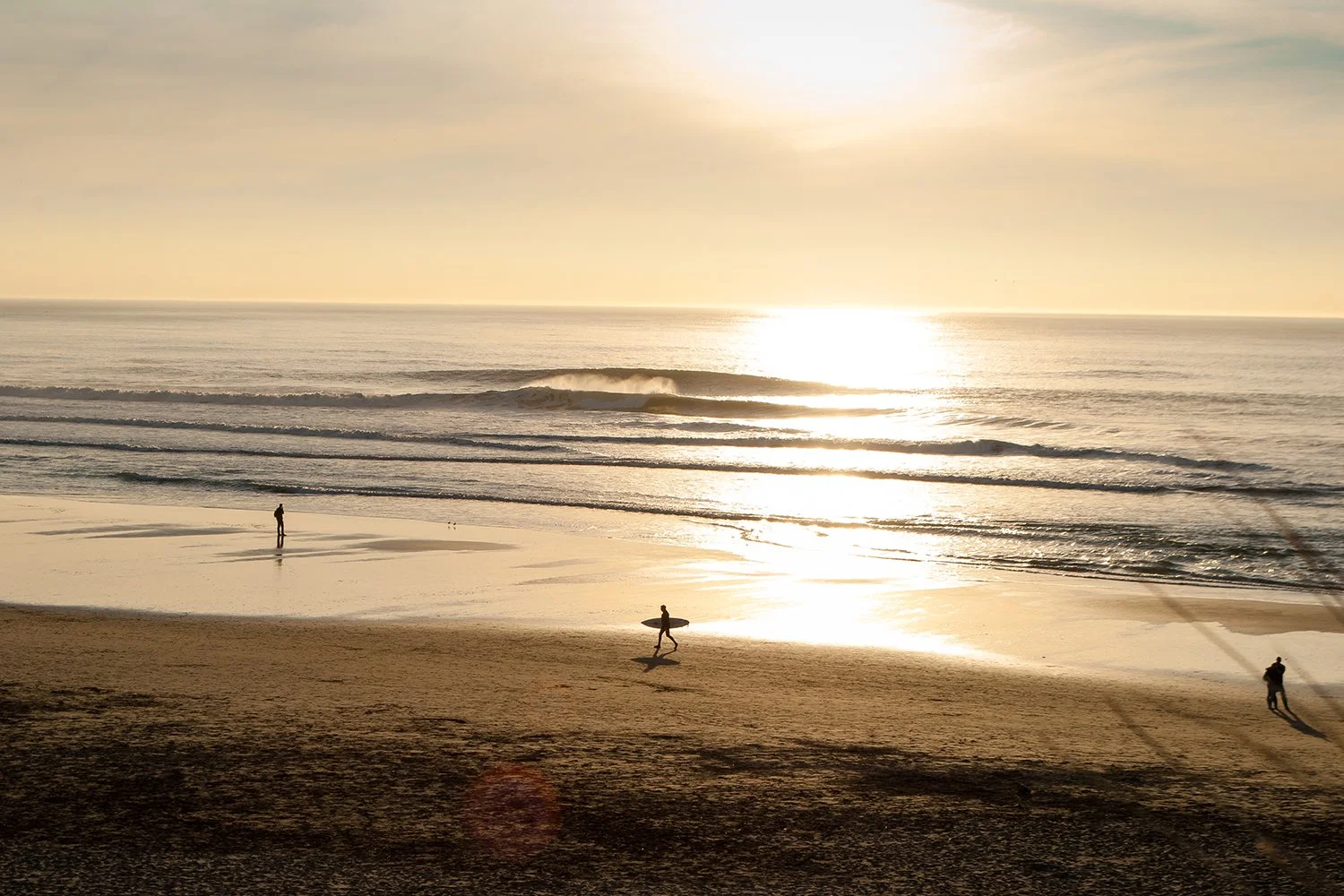 Sunset on a beach with three surfers walking along the sand and carrying surfboards, calm ocean waves near shore, warm golden sky.
