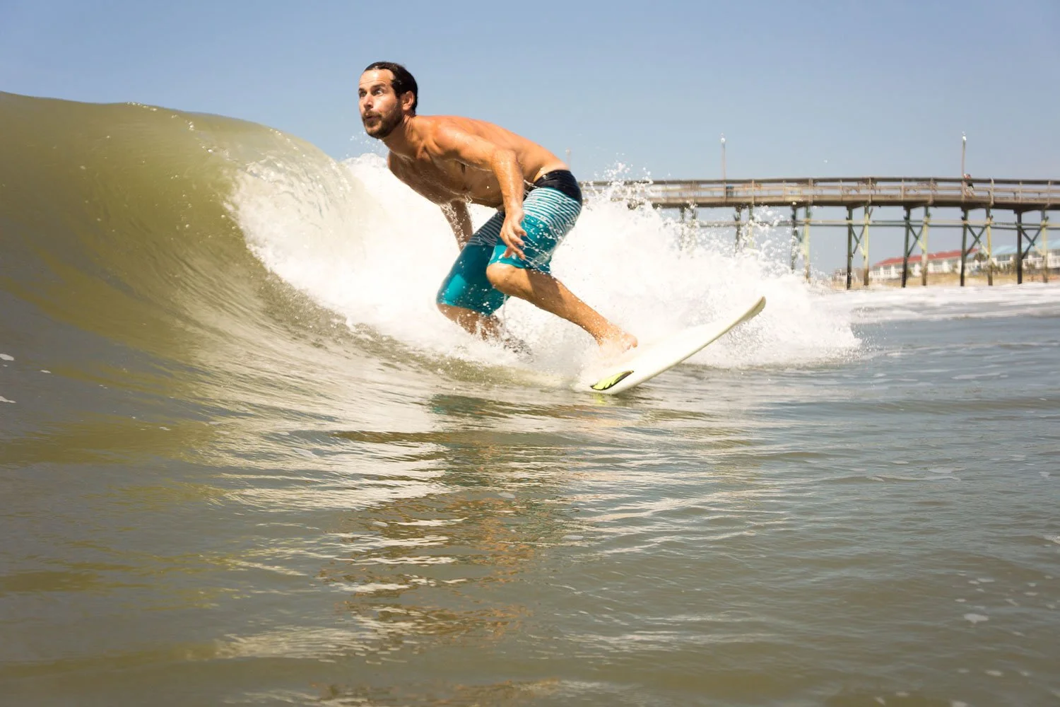 Man surfing on a wave at the beach with a pier in the background on a clear day.