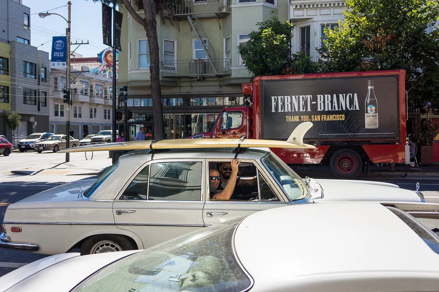 People in a white vintage car parked on a city street with a surfboard on top, and a red delivery truck in the background with an advertisement for Fernet-Branca thanking San Francisco.