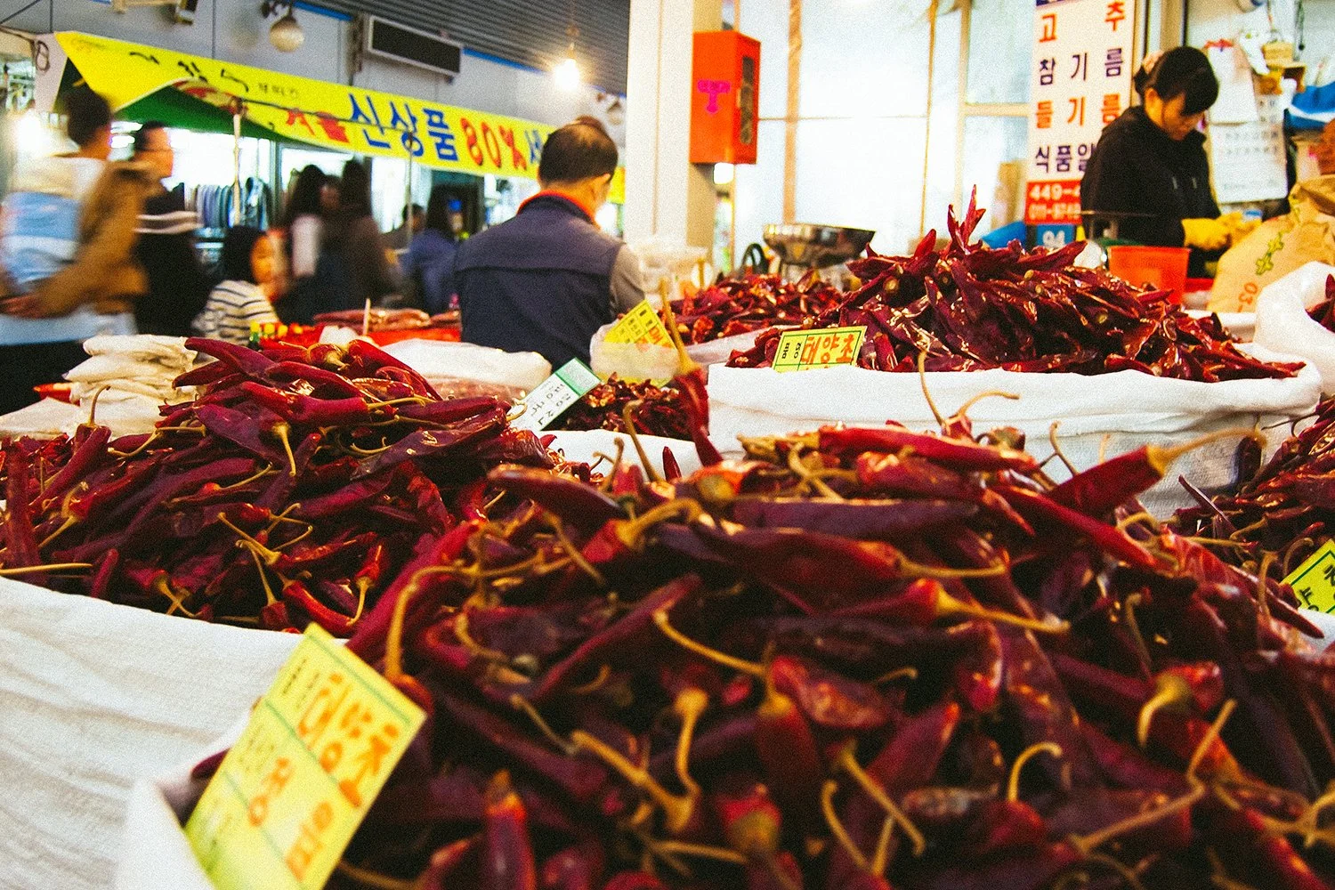 A market stall with piles of dried red chili peppers for sale, with shoppers and vendors in the background.