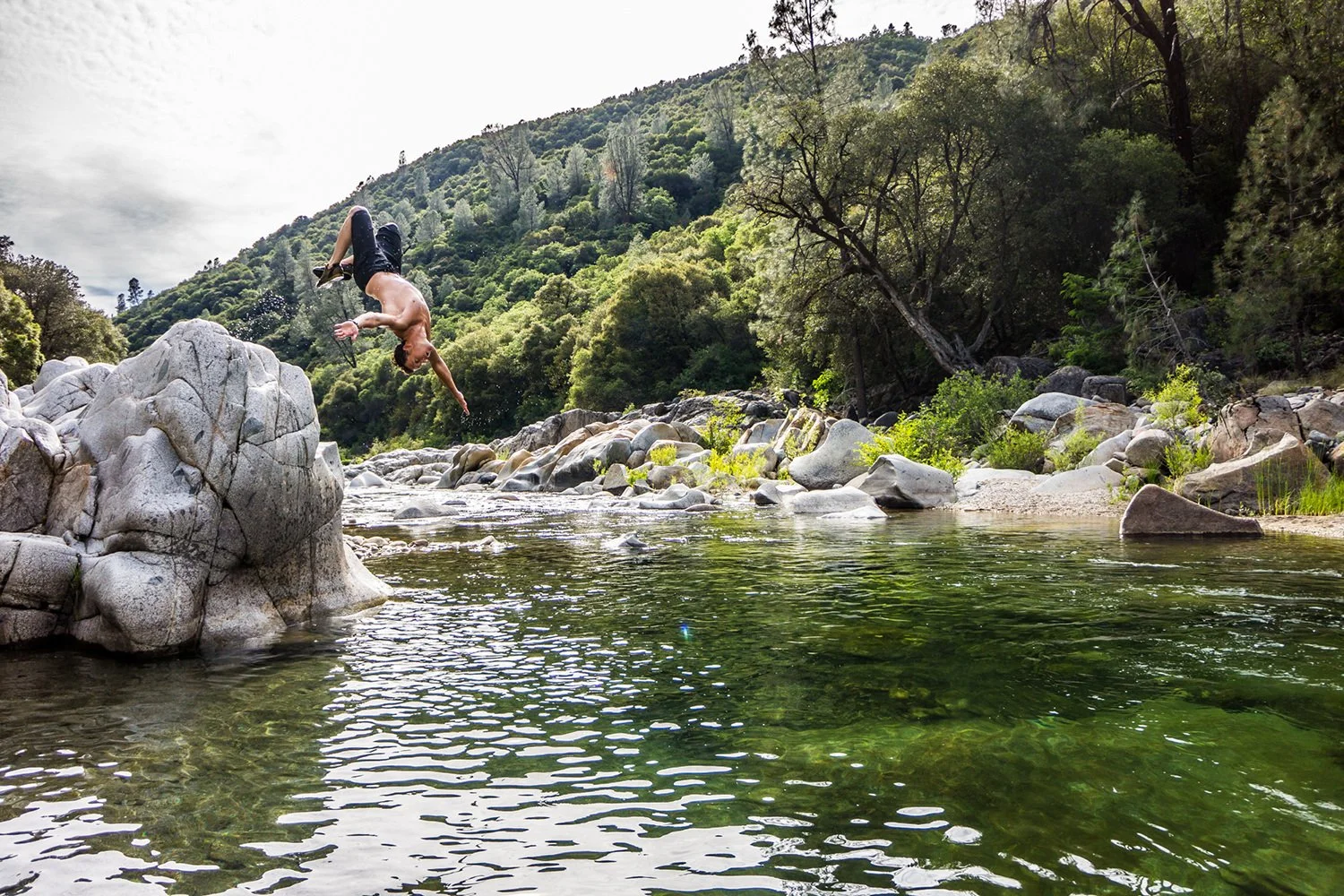 A man jumping into a river surrounded by rocks and lush green trees on a sunny day.