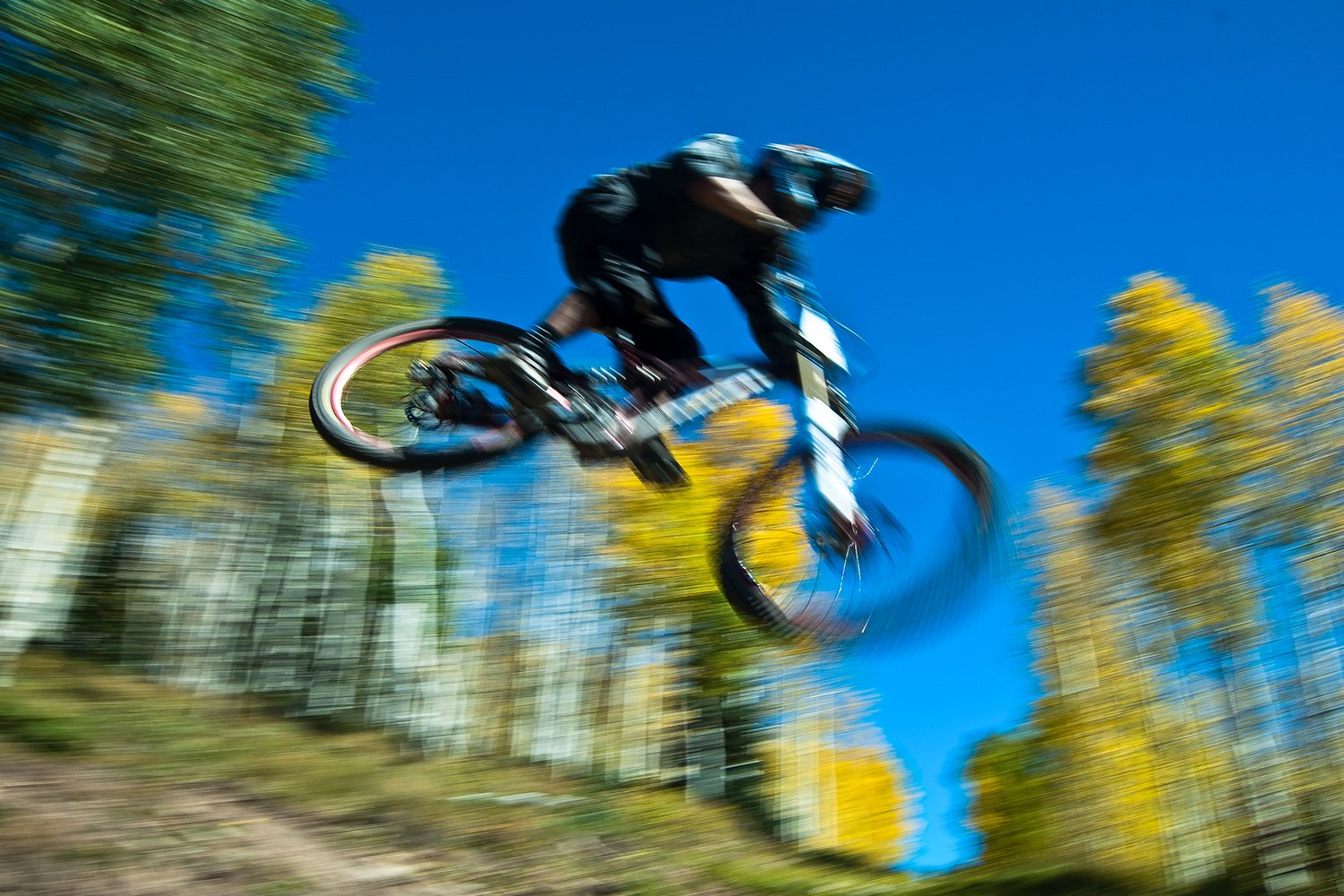 A mountain biker in black riding gear launching into a jump on a trail, captured in motion with a blurred background of autumn trees and clear blue sky.