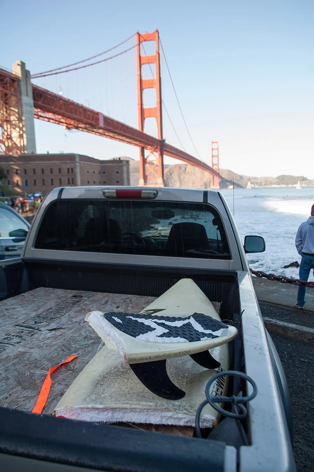 A pickup truck with a surfboard and a wetsuit in the truck bed, with the Golden Gate Bridge in the background on a clear day.