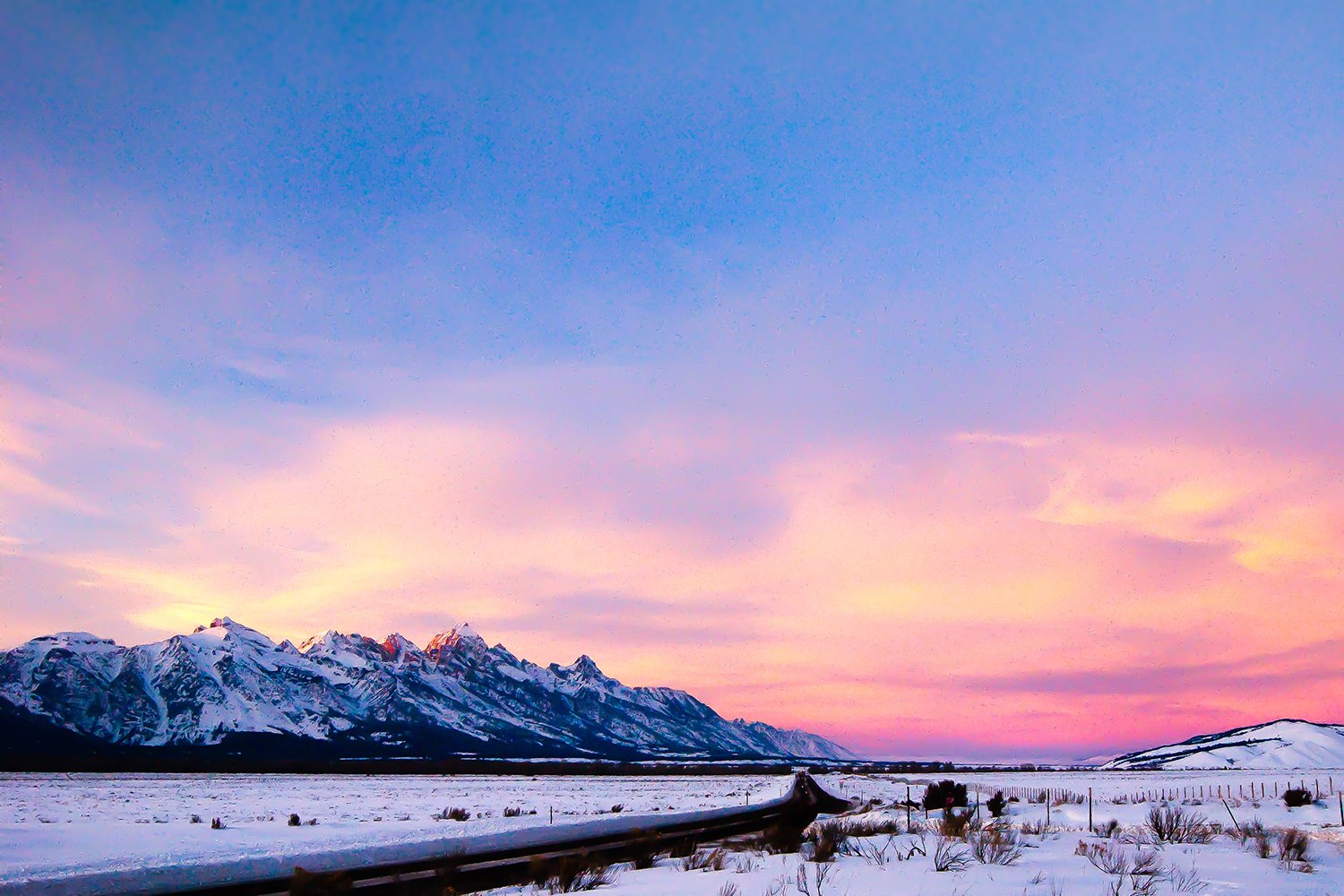 Snow-covered fields with mountains in the background and a colorful sunset sky with pink, purple, and blue hues.