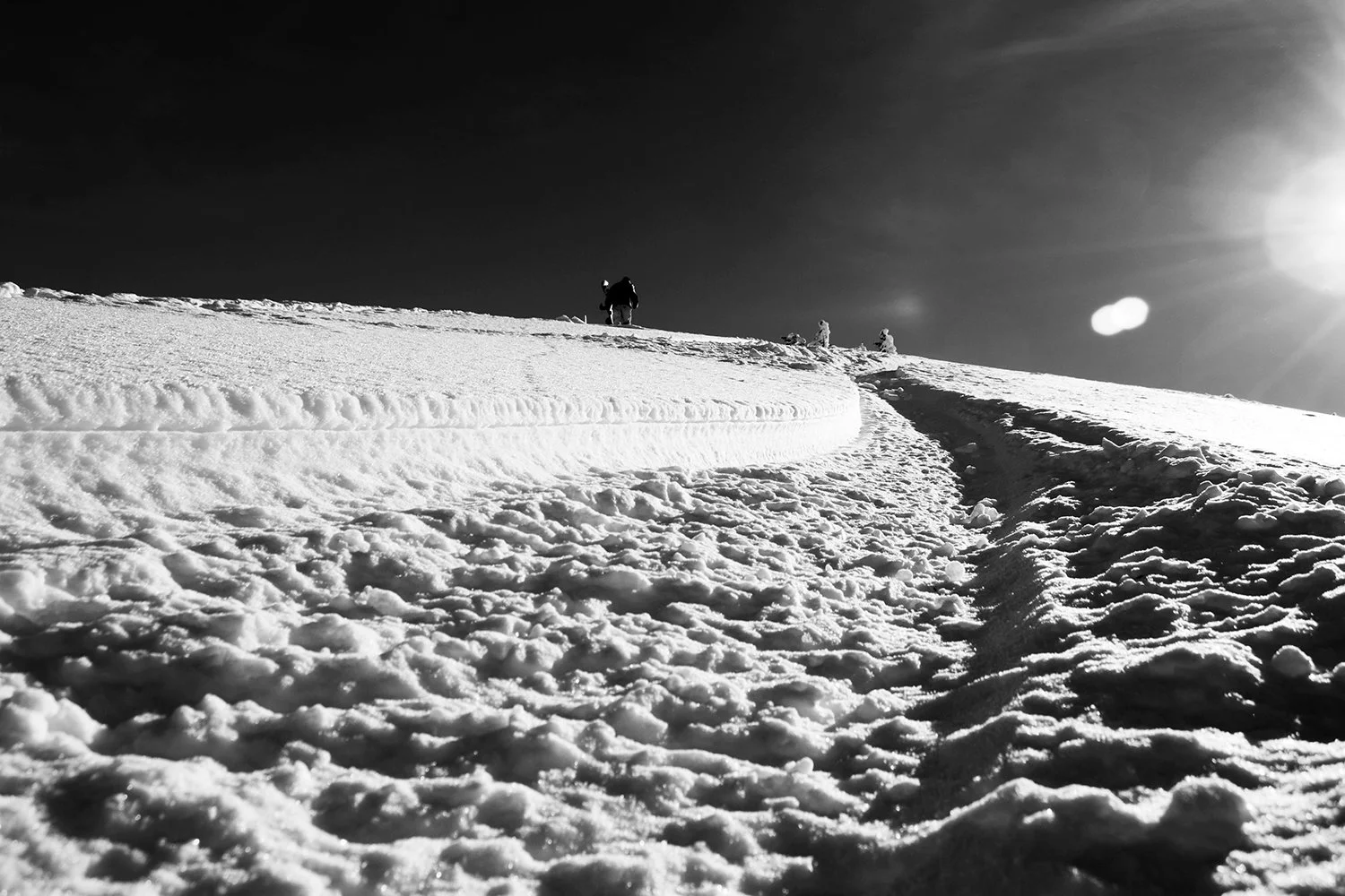 Snowy mountain slope with a trail leading to the top, where a few people are standing. The sky is clear and the sun is shining brightly on the right side.