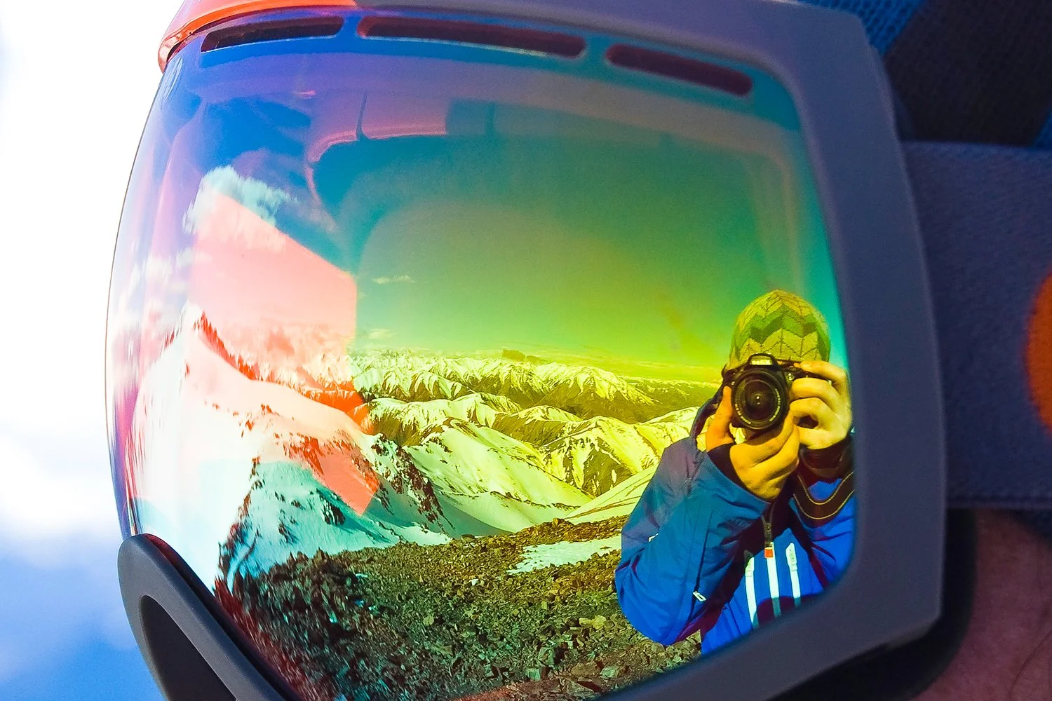 Reflection of a person taking a photograph with a camera, wearing a blue jacket and a patterned hat, in a mountain landscape with snow-capped peaks, displayed in a tinted visor or mirror.