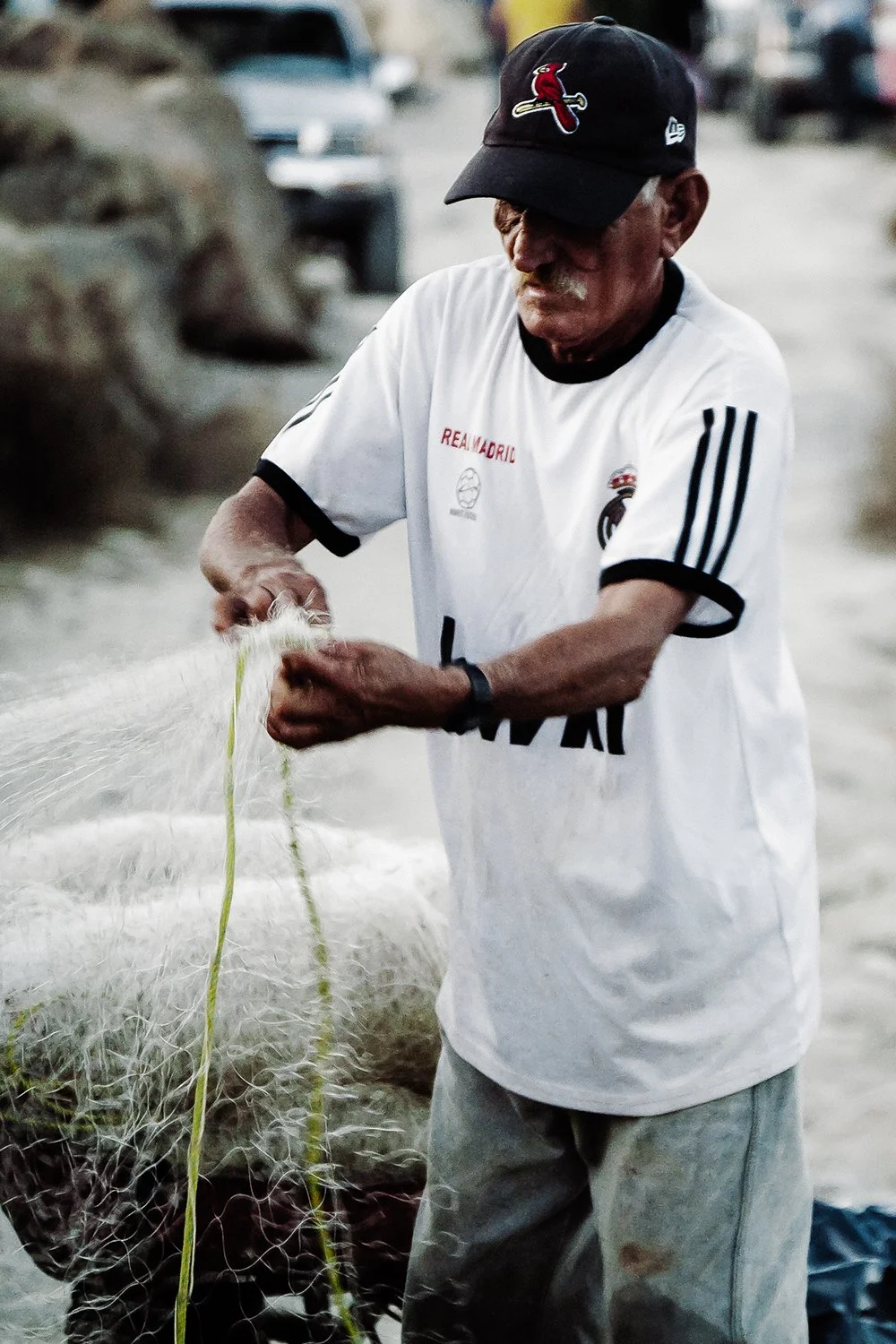 An elderly man wearing a black baseball cap with a red cardinals logo, a white soccer jersey with black trim, and gray pants, standing outdoors handling fishing net.