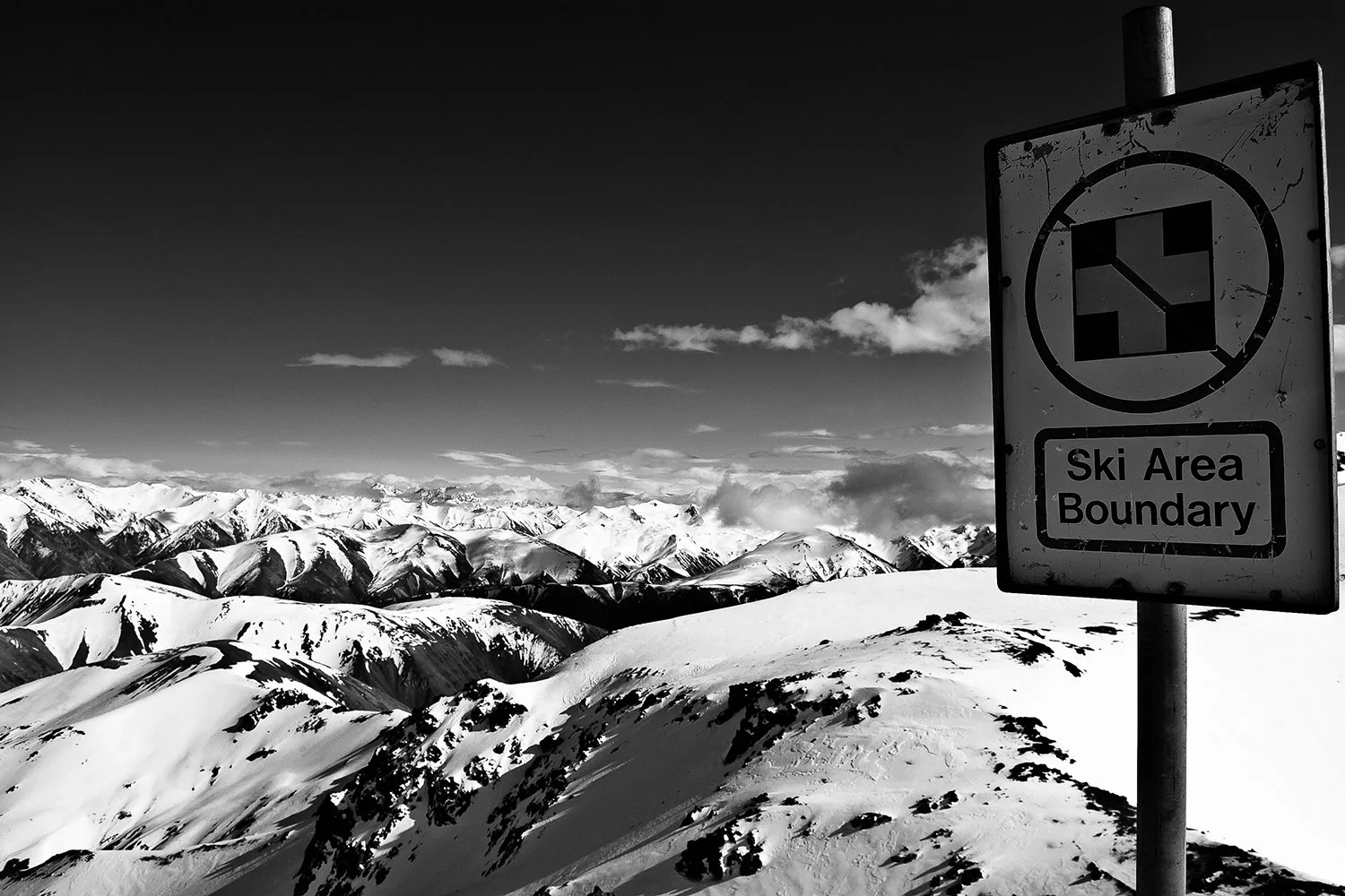 Snow-covered mountain range with a sign indicating the boundary of a ski area in black and white.