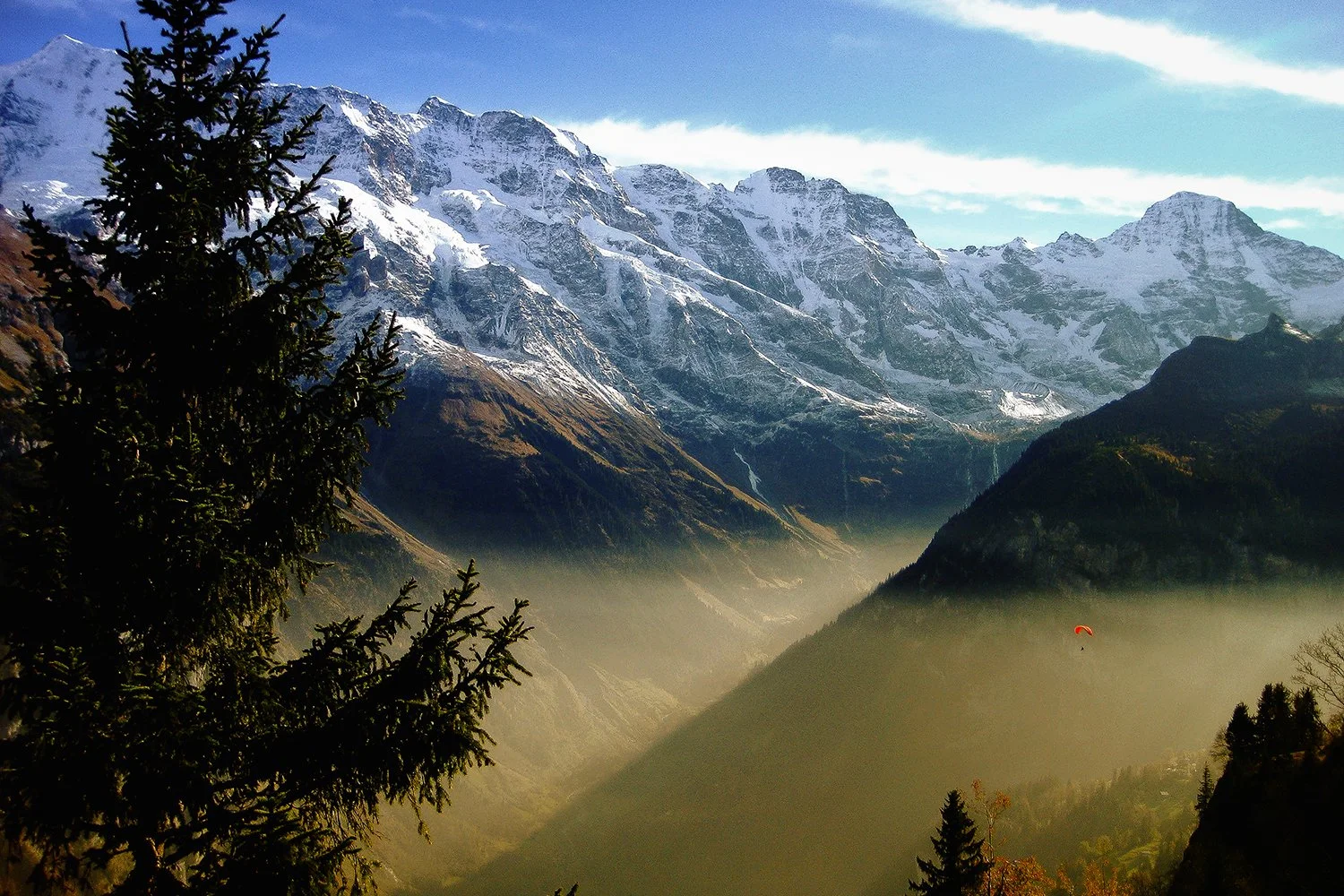 Snow-capped mountains with green valleys and a paraglider flying over the misty landscape during daytime.