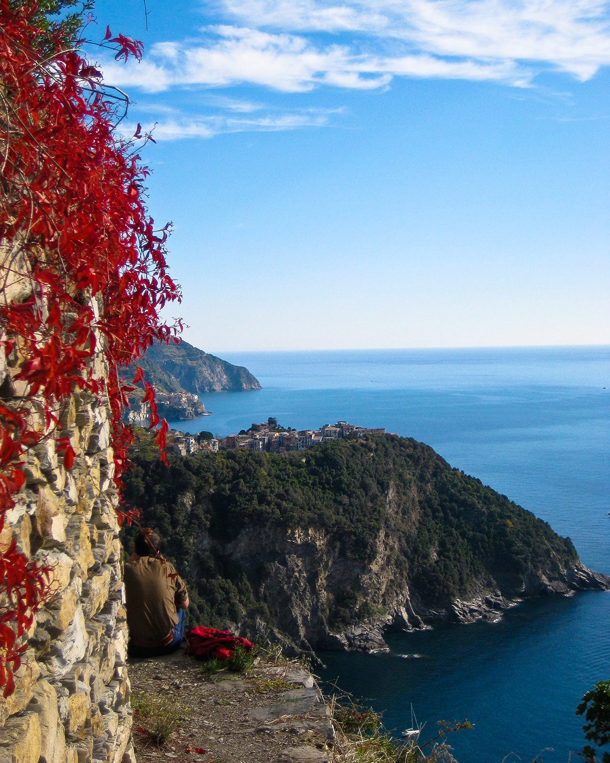 Person sitting on a ledge overlooking coastal cliffs and the ocean, with a red vine-covered wall on the left side and a small town in the distance under a blue sky.