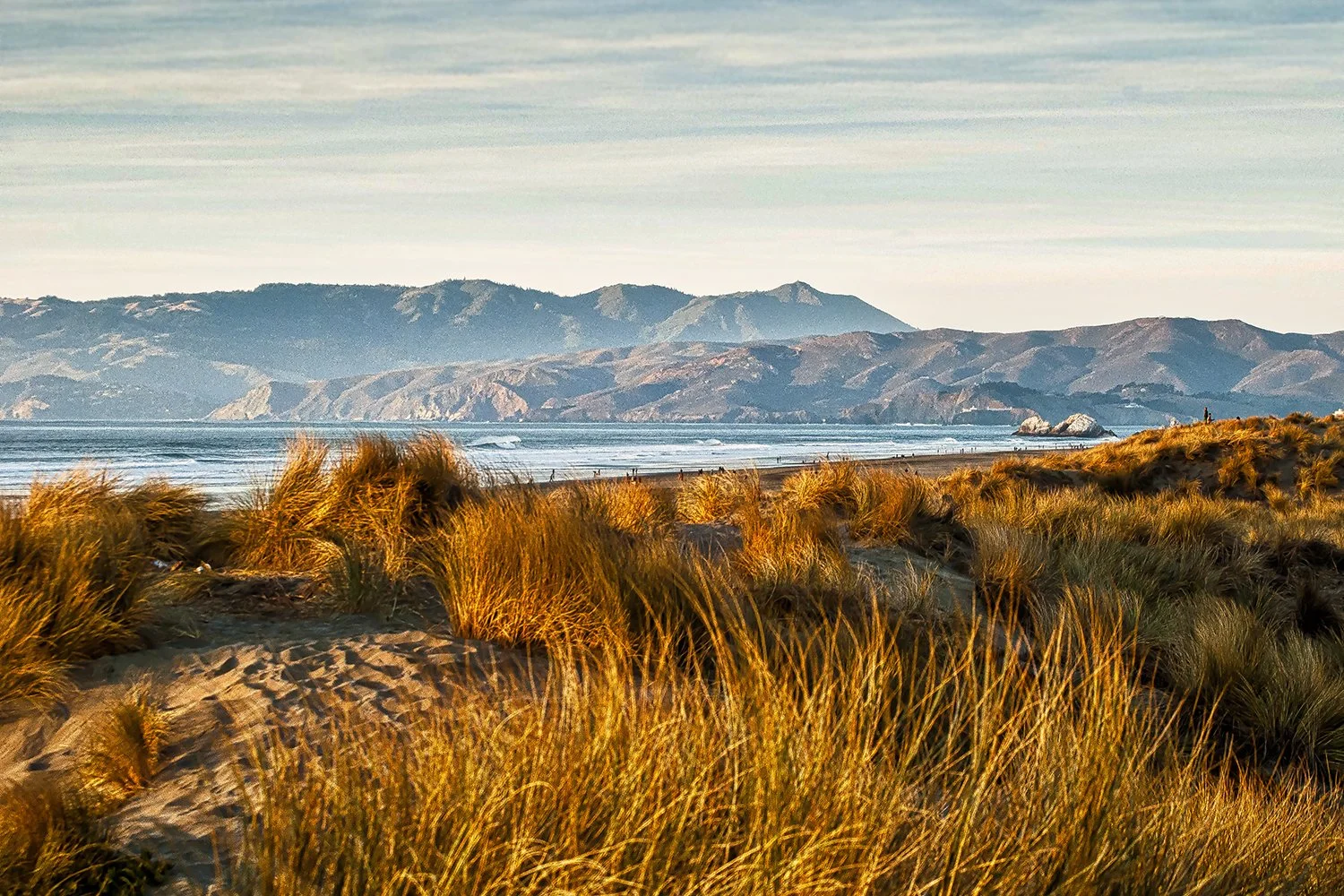 A coastal landscape with sandy dunes and tall grasses in the foreground, the ocean with gentle waves in the middle ground, and mountain ranges in the background under a partly cloudy sky.