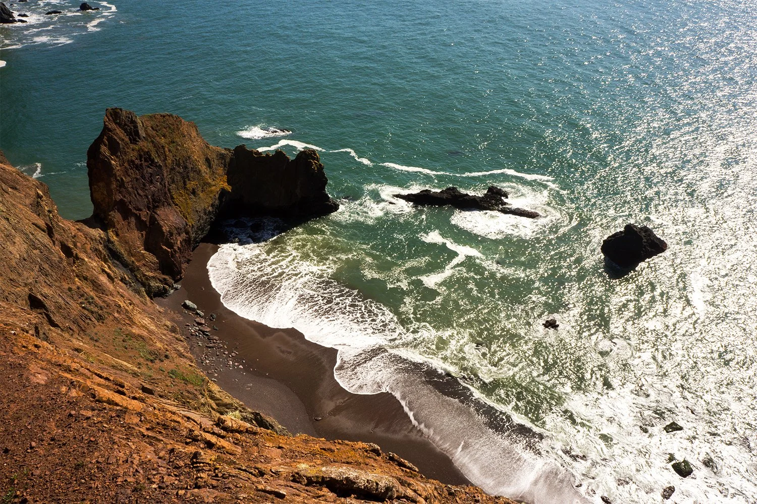 Aerial view of a rugged coastline with large rocks and cliffs, black sandy beach, and the ocean with white foam surf reflecting sunlight.
