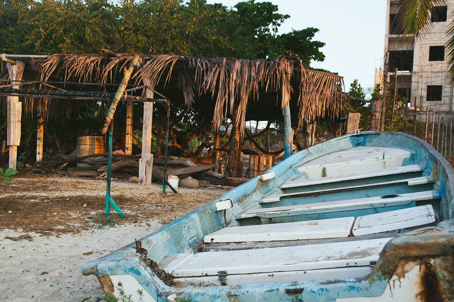 An abandoned, weathered boat on a sandy beach with a rustic hut made of wooden poles and dried palm leaves in the background, surrounded by lush green trees and an unfinished building.