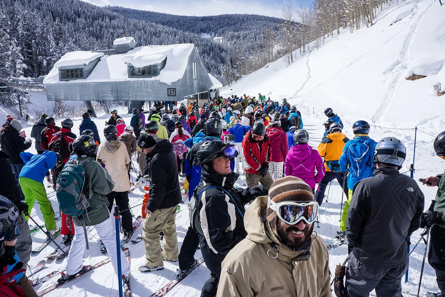 Crowd of skiers and snowboarders waiting in line at a snowy ski resort with a chairlift in the background.