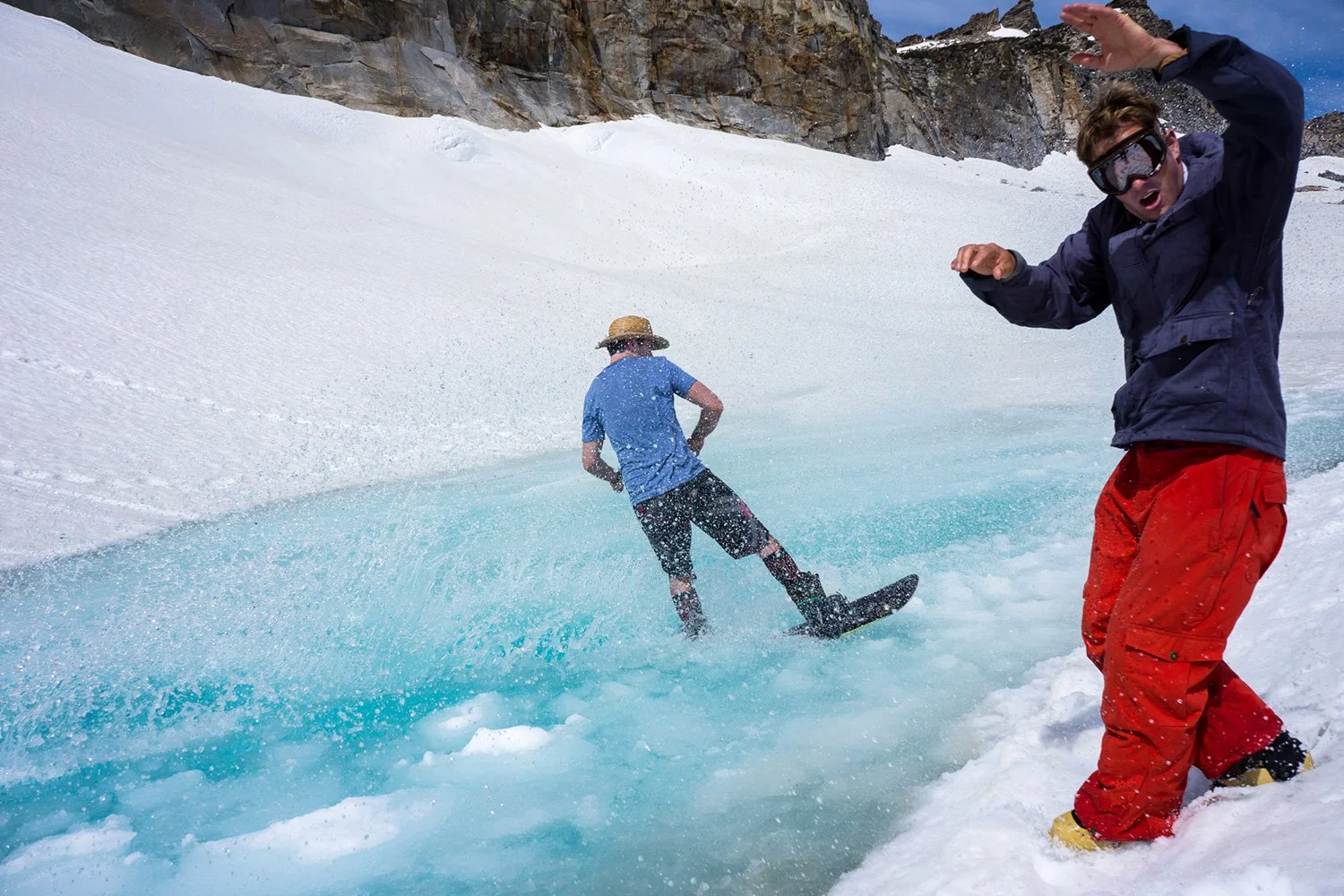 Two men standing on snowy ice near a small meltwater pond with a mountain rocky backdrop. One man is skiing into the pond while the other is standing nearby wearing sunglasses and red pants, raising hand gesture.