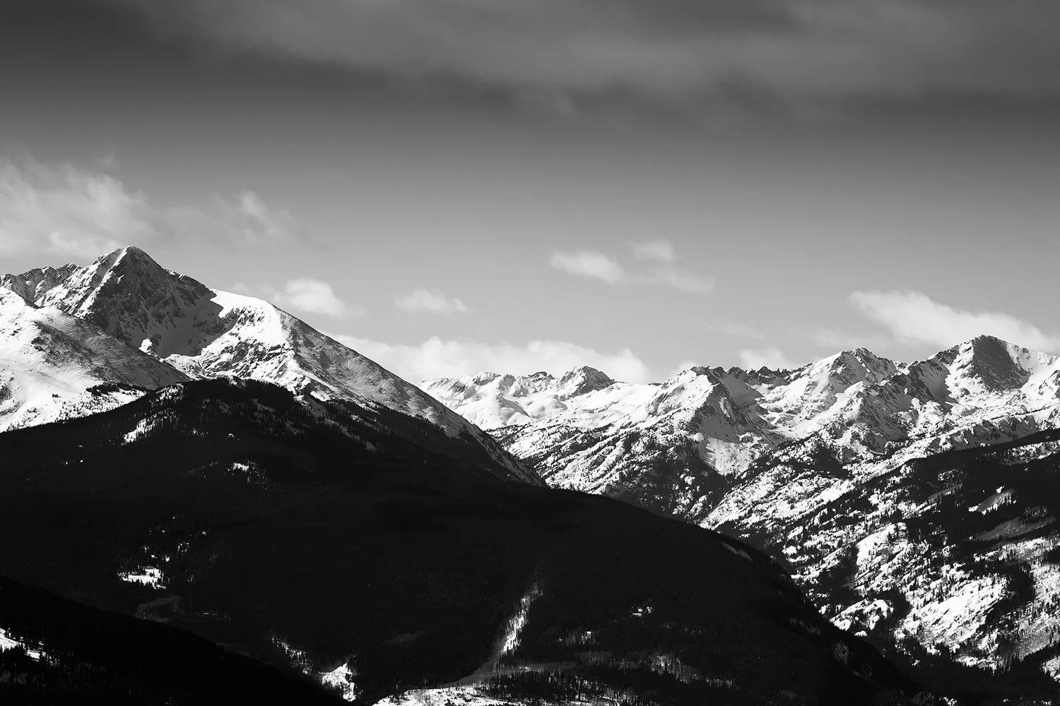 Snow-covered mountains with dark forests at their base, under a partly cloudy sky.