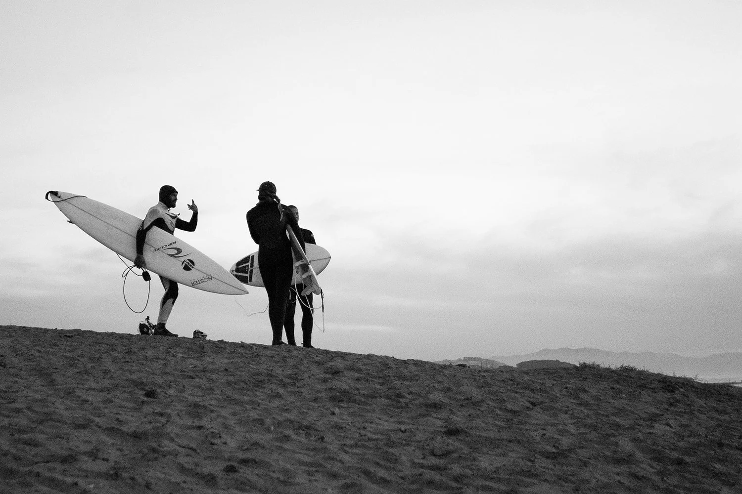 Three surfers stand on a sandy beach with their surfboards, engaging in conversation in a black and white photo, with a distant mountain range visible in the background.