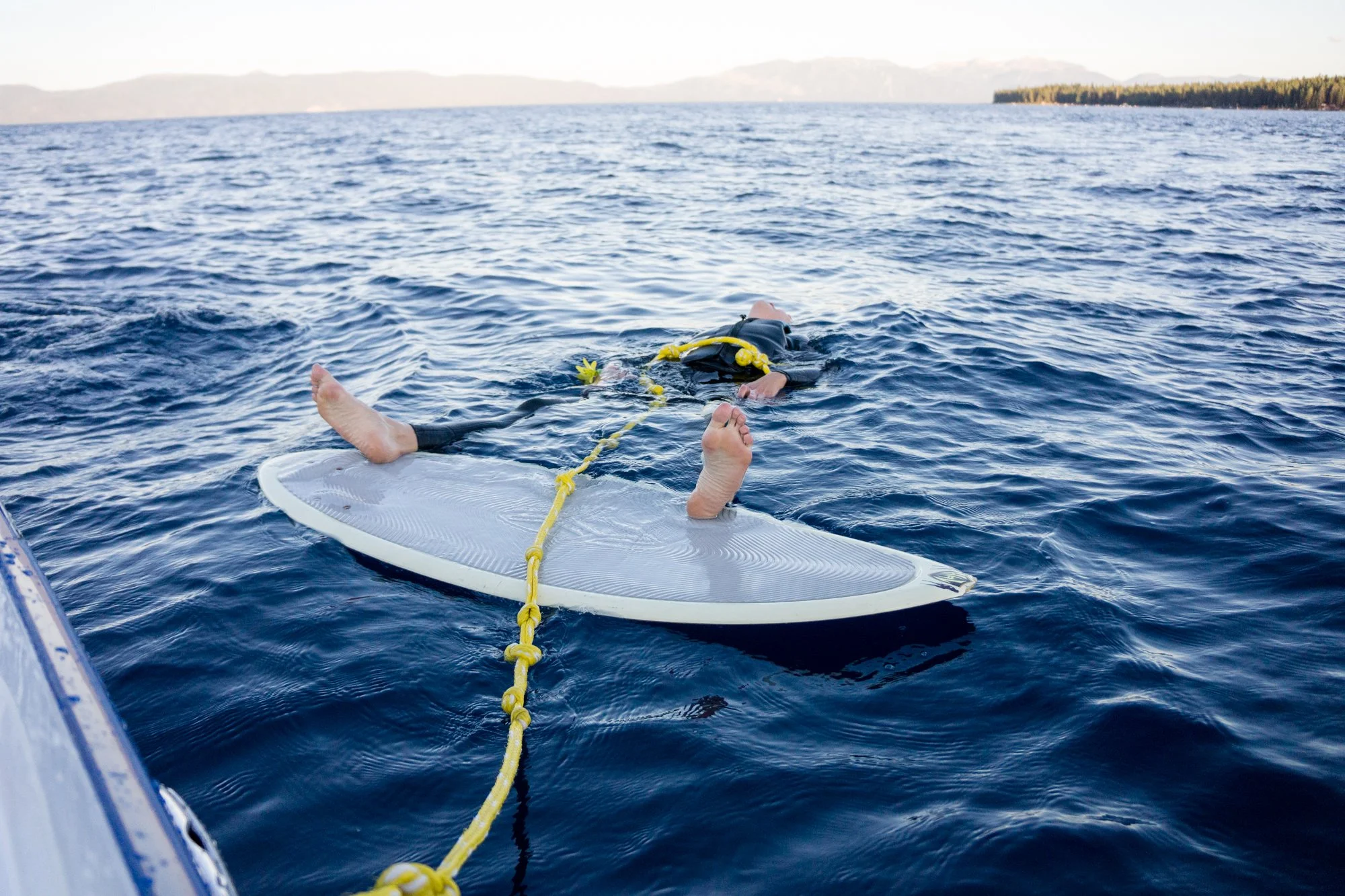 A person floating on a paddleboard in open water, holding onto a yellow rope attached to a rescue swimmer in a wetsuit. The swimmer is face down in the water near the paddleboard, with a life jacket and flippers. A distant shoreline and mountains are
