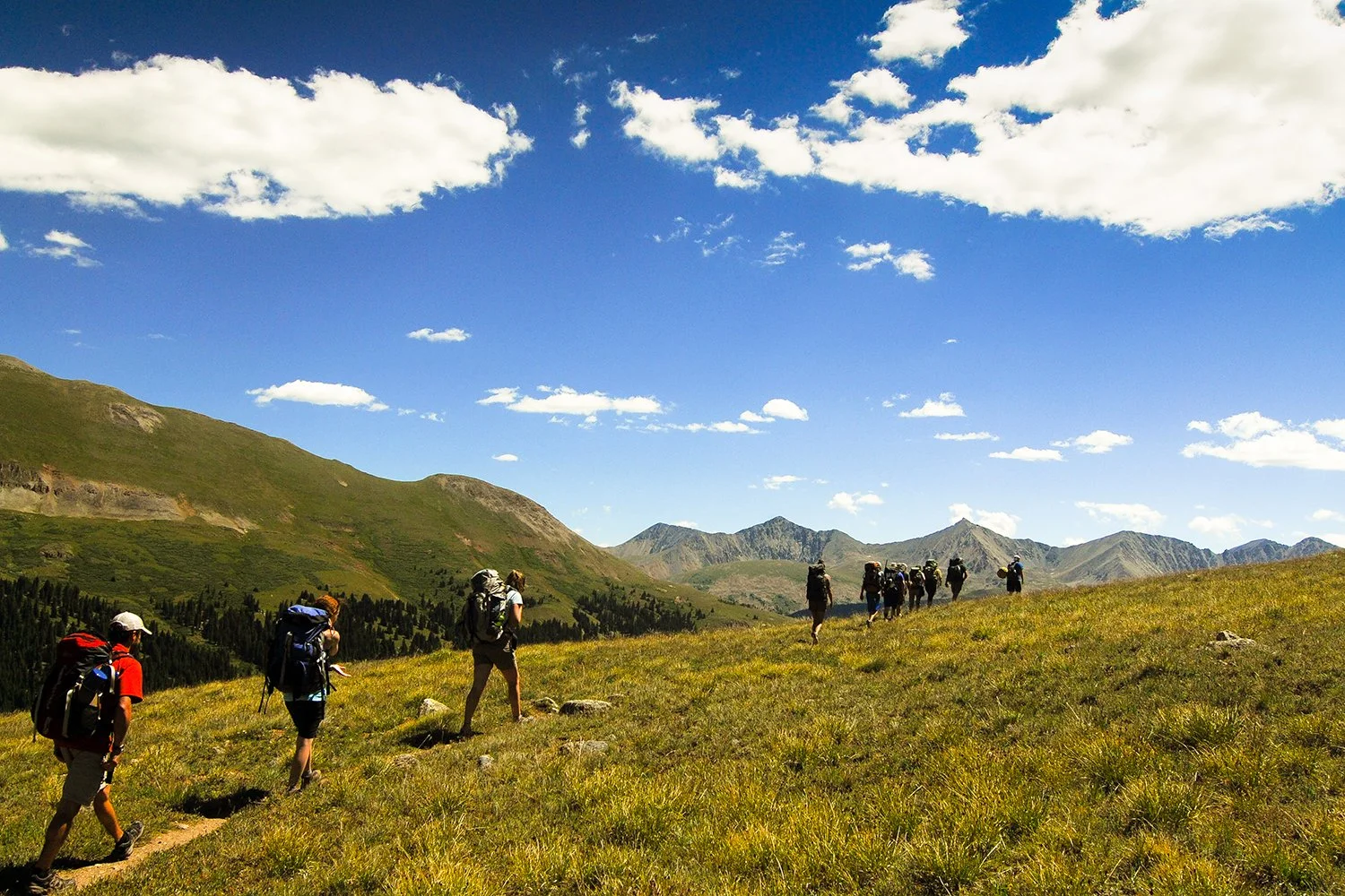 A group of hikers walking through a grassy mountain landscape under a blue sky with scattered clouds.