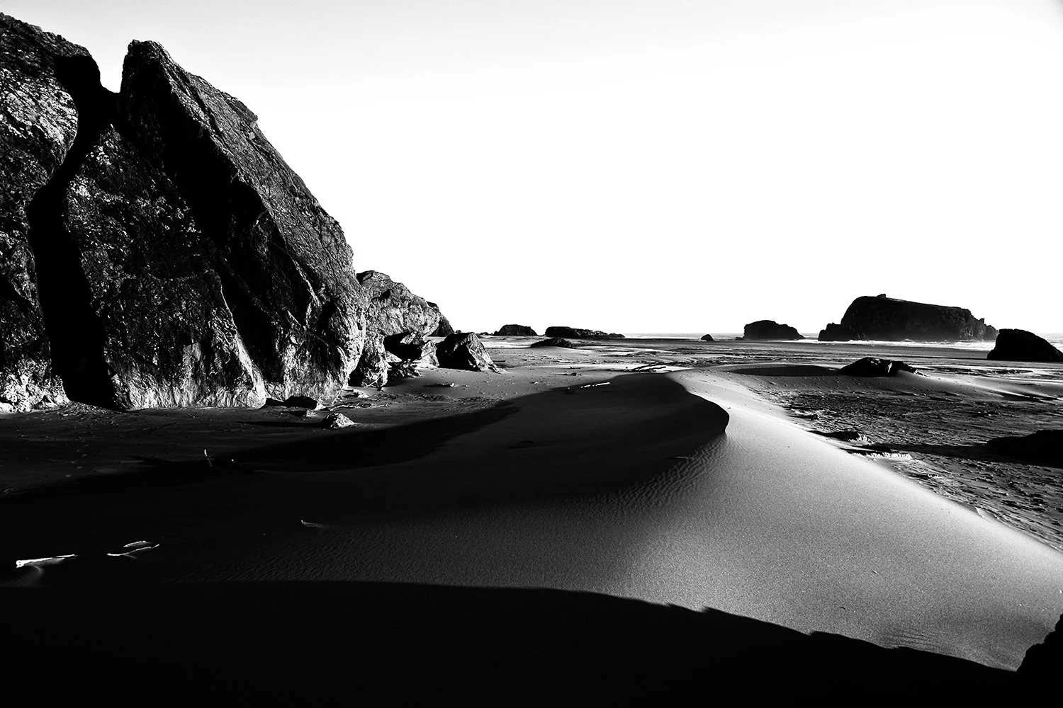 Black and white photo of a beach with large rocks and sand dunes, ocean in the background, and shadows cast on the landscape.