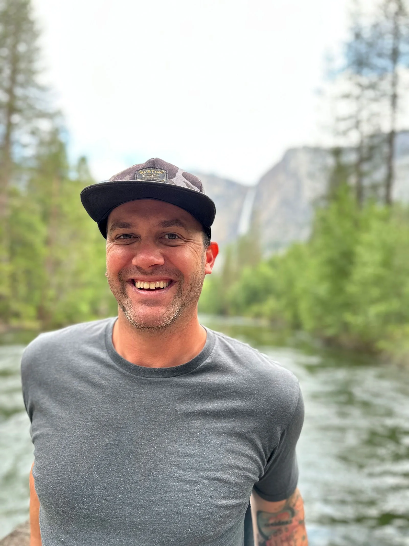 A smiling man wearing a gray t-shirt and a camouflage cap with a Burton logo, outdoors in front of a river with trees and a waterfall in the background.