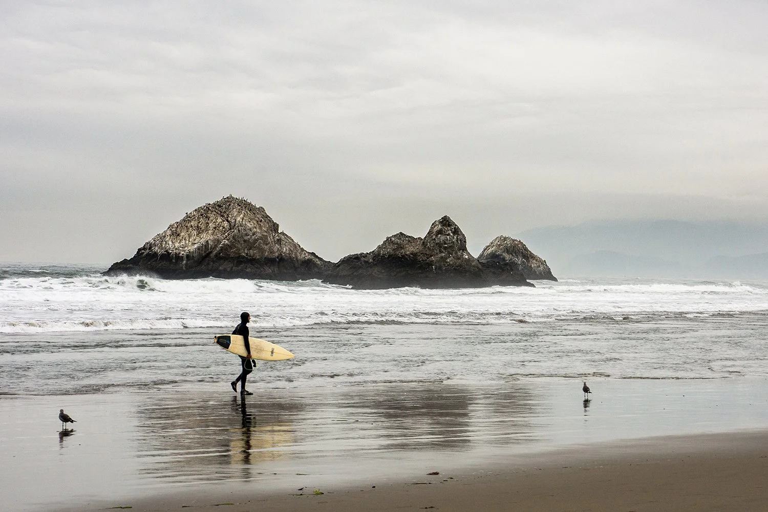 Person walking on the beach with a surfboard, near the water, with rocks and waves in the background.