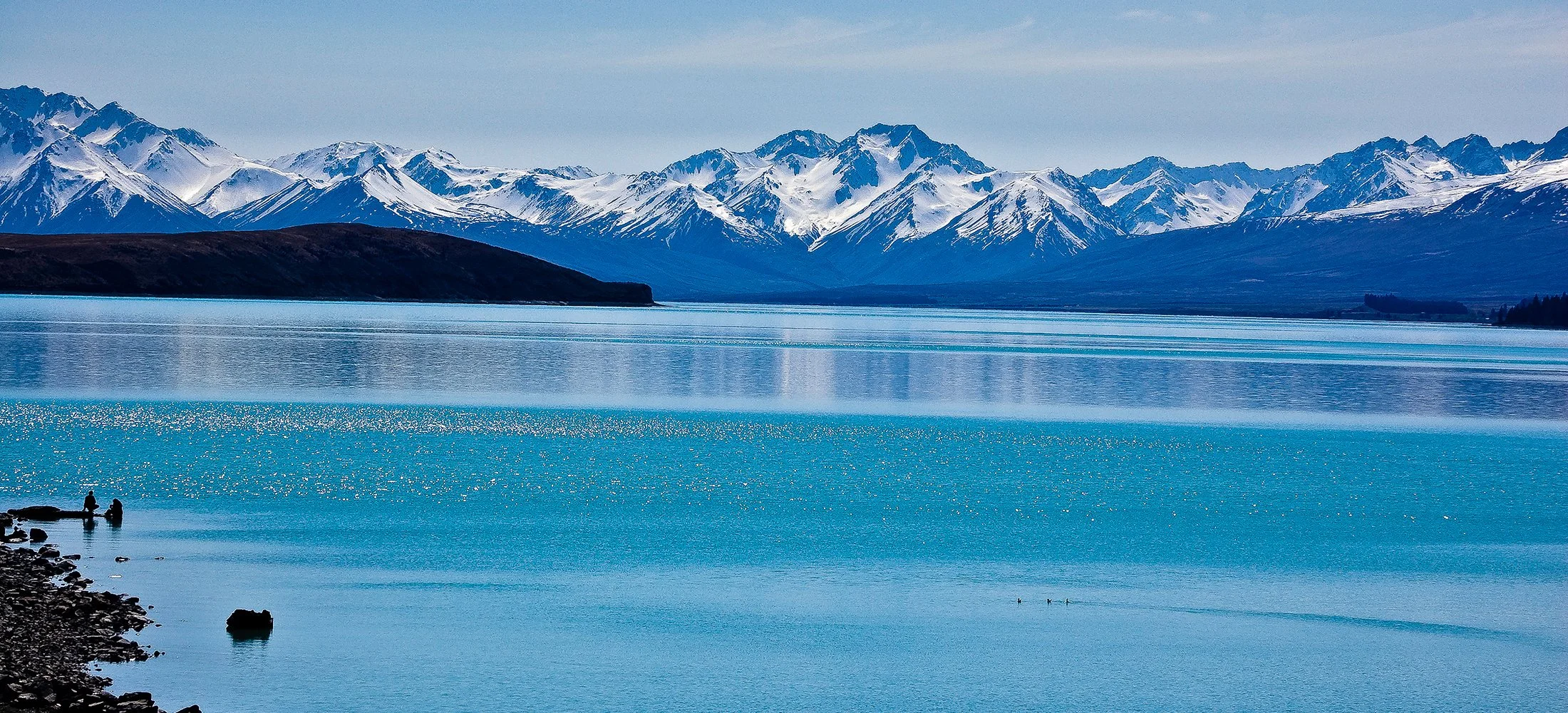 A body of water with two people fishing on the rocky shoreline, snow-capped mountains in the background, and a calm, blue sky overhead.