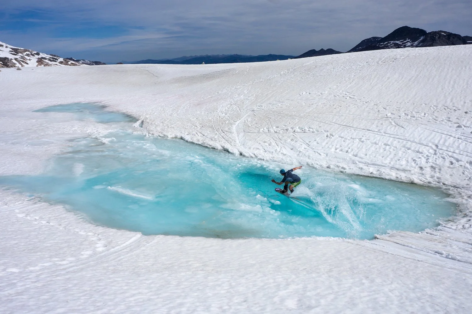 A person snowboarding on a snow-covered slope near a patch of blue melting snow or ice in a mountain landscape.