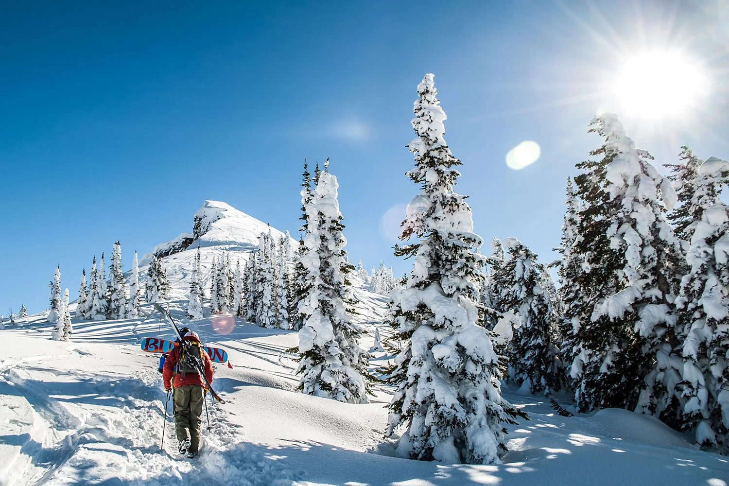 A person hiking in a snowy mountain landscape with snow-covered trees, a mountain peak in the background, and the sun shining brightly in a clear blue sky.