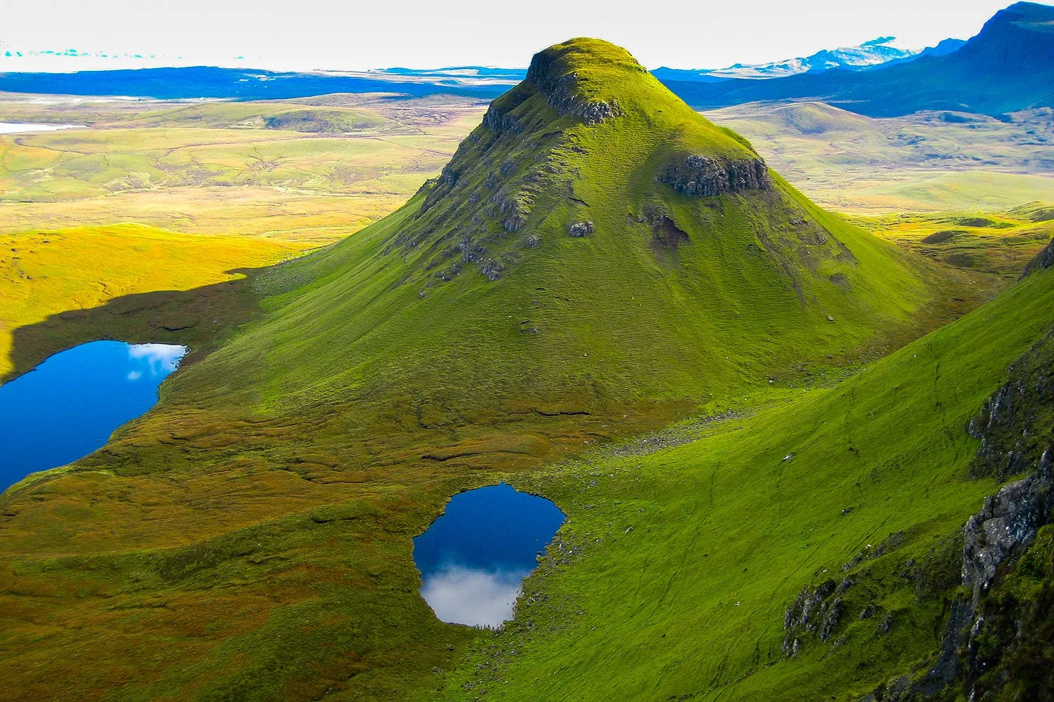A lush green mountain with a flat top, surrounded by two small lakes with blue reflections, under a partly cloudy sky.