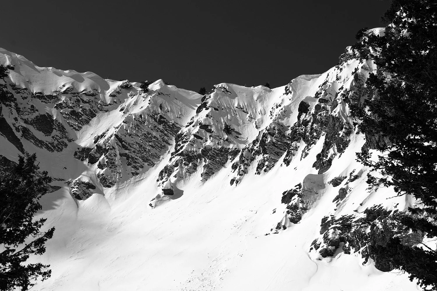 Snow-covered mountain range with steep slopes, rocky outcroppings, and sparse trees in the foreground against a dark sky.