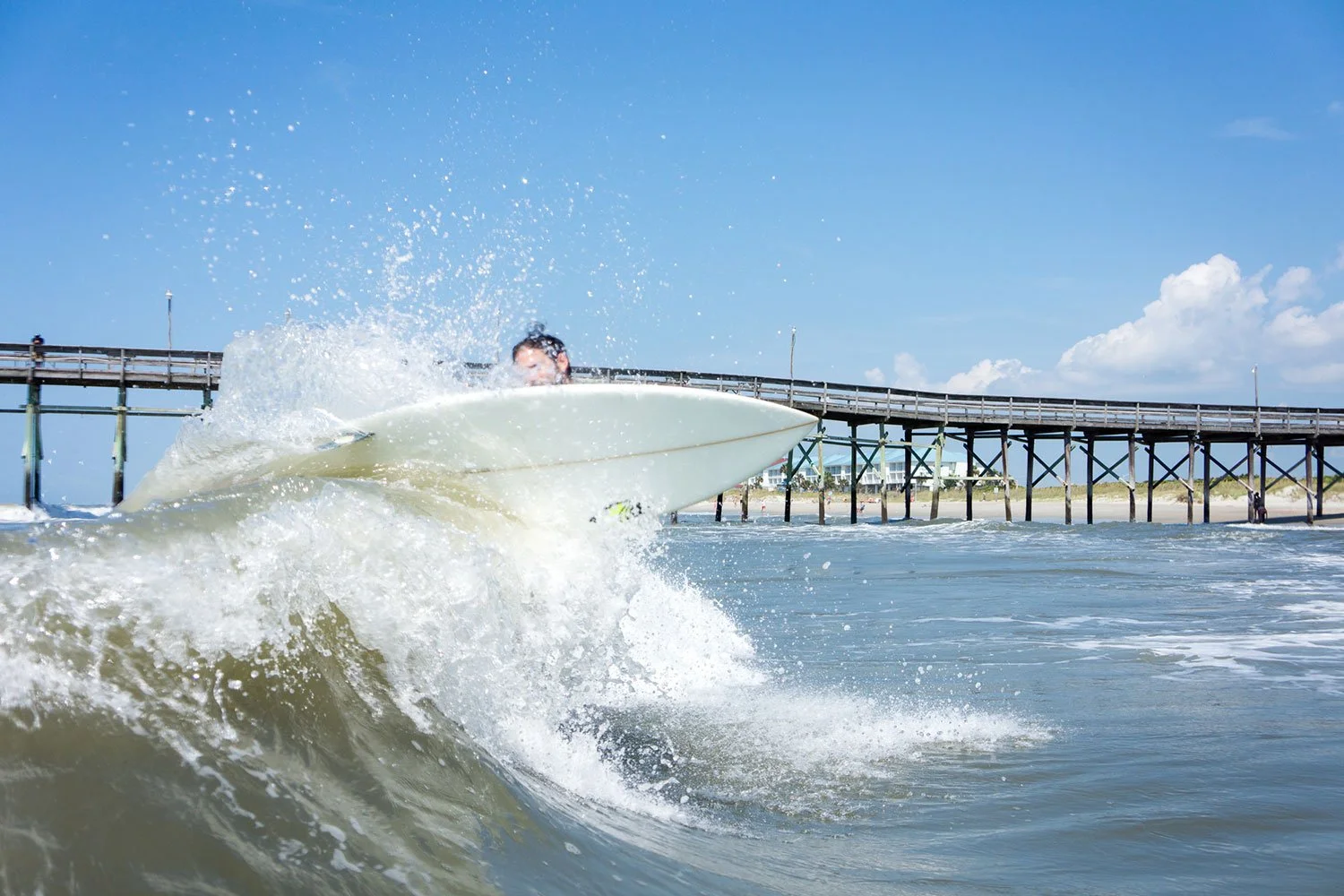 A person surfing on a wave near a pier on a sunny day with a clear blue sky.