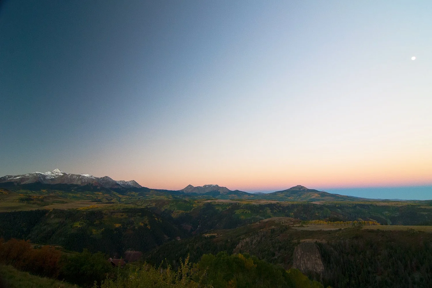 A landscape view of mountains at dusk with snow-capped peaks, rolling hills, and green forests under a sky transitioning from sunrise to twilight, with a small bright moon visible.