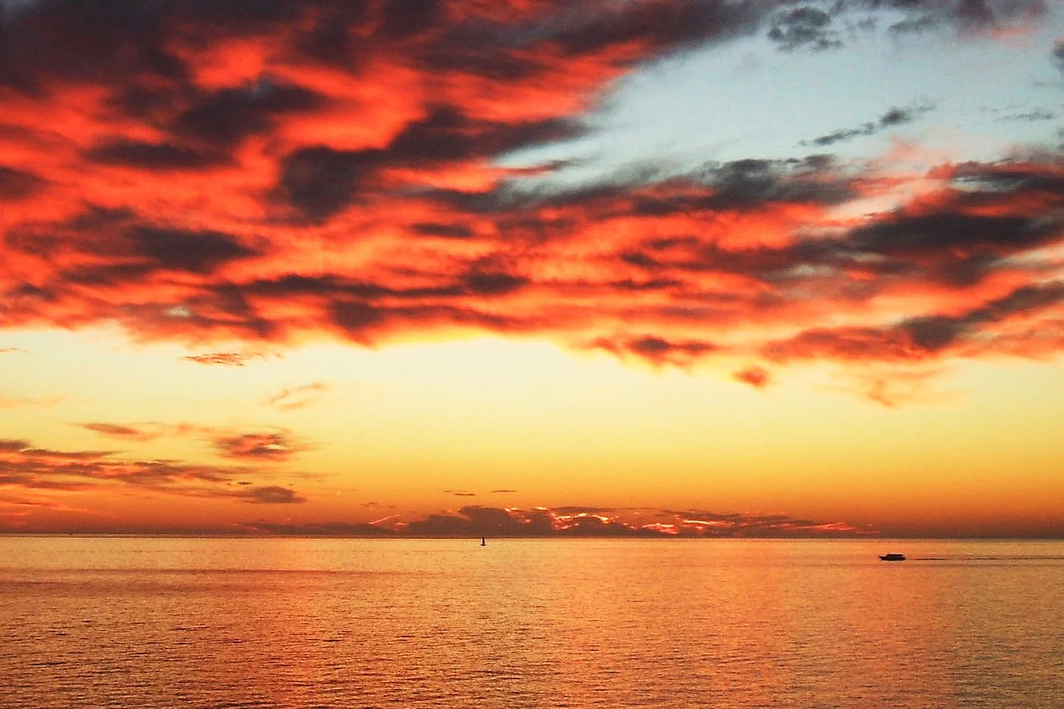 A sunset over the ocean with a colorful sky filled with red, orange, and pink clouds, and a boat on the water.