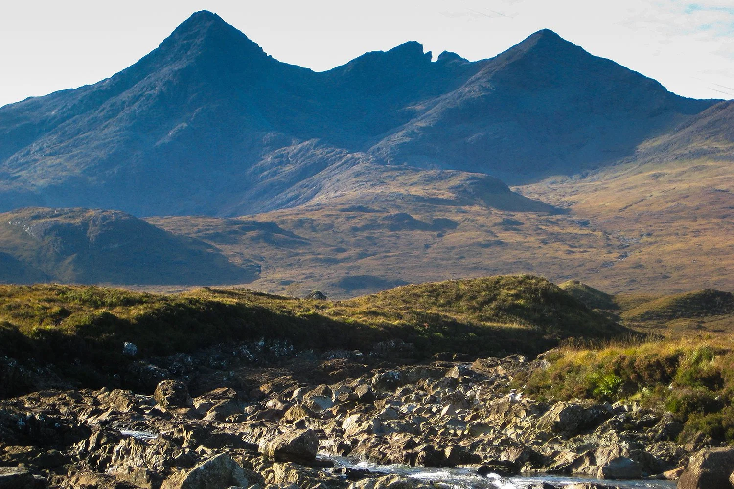 Mountain landscape with rugged peaks in the background and grassy hills in the foreground, with a small rocky stream flowing through the terrain.