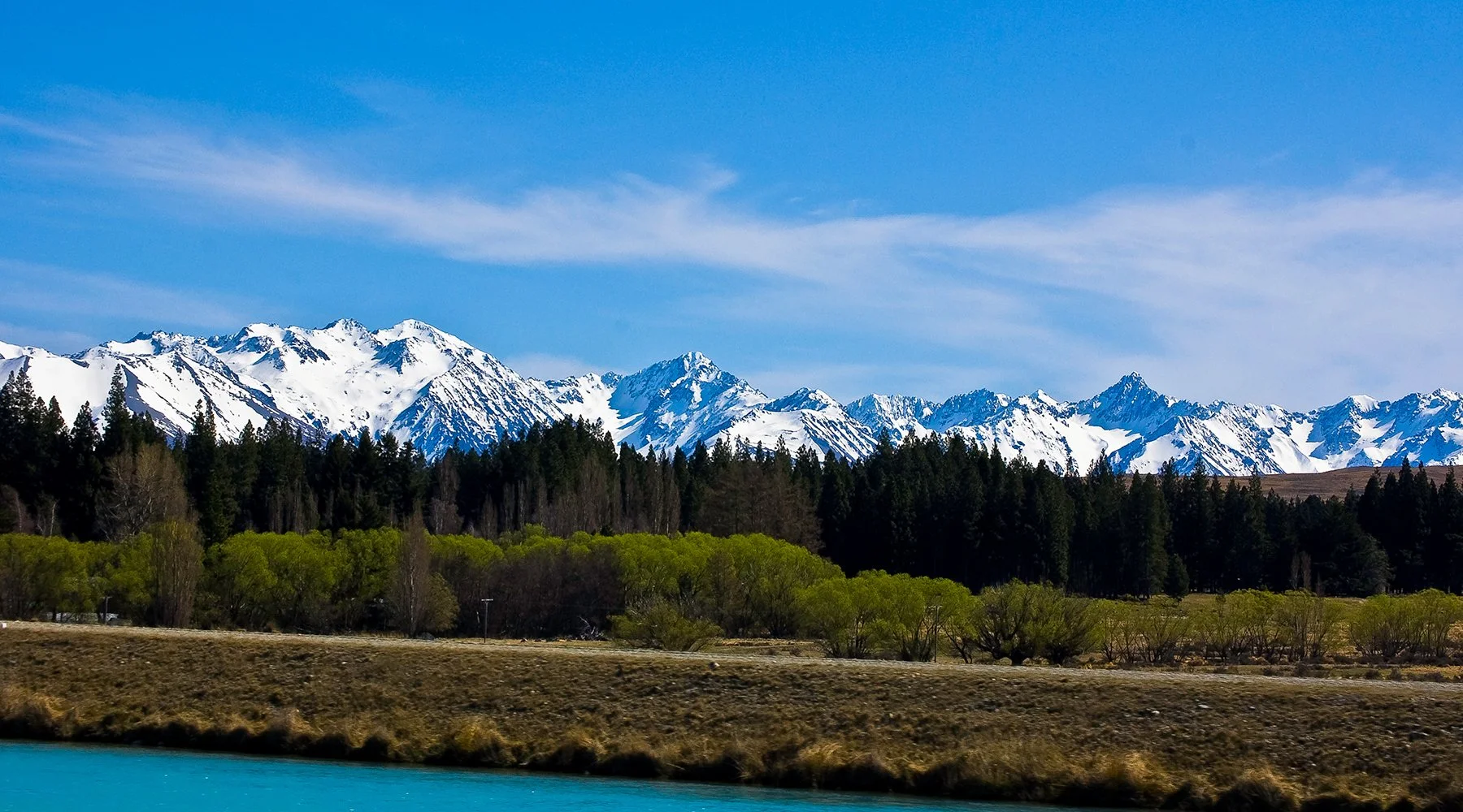 Snow-capped mountains in the distance with a forest of green trees and a body of blue water in the foreground.