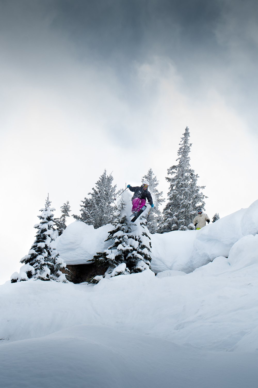 A person in winter gear snowboarding off a snow-covered ledge in a snowy mountain landscape with trees.