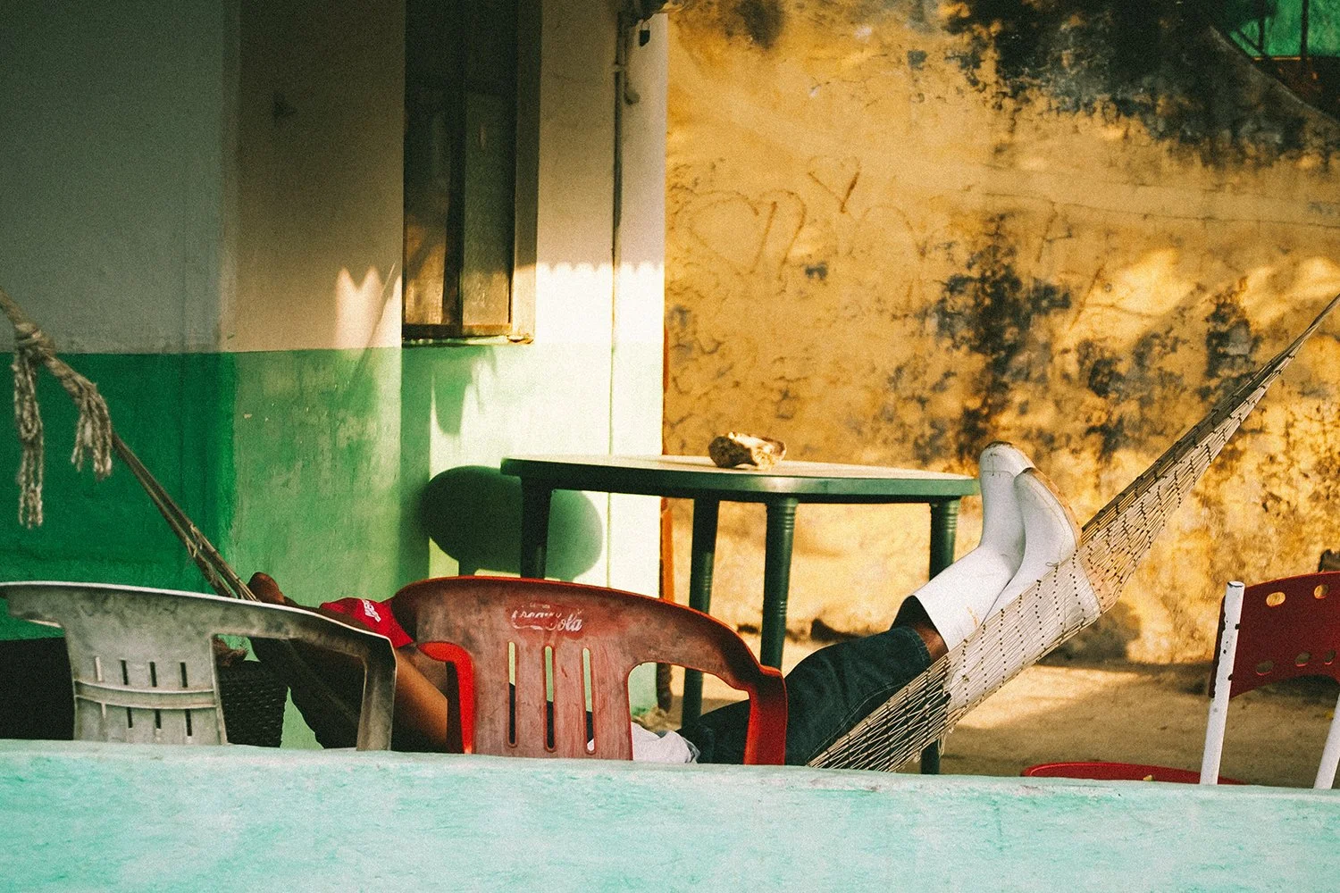 Person relaxing in a hammock with their feet up, surrounded by plastic chairs and a table outdoors, in front of a weathered wall with graffiti.
