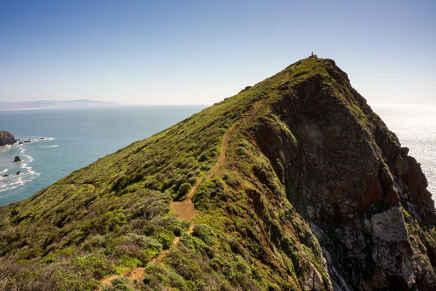 A grassy trail on a mountain ridge overlooking the ocean, with a person standing at the peak.