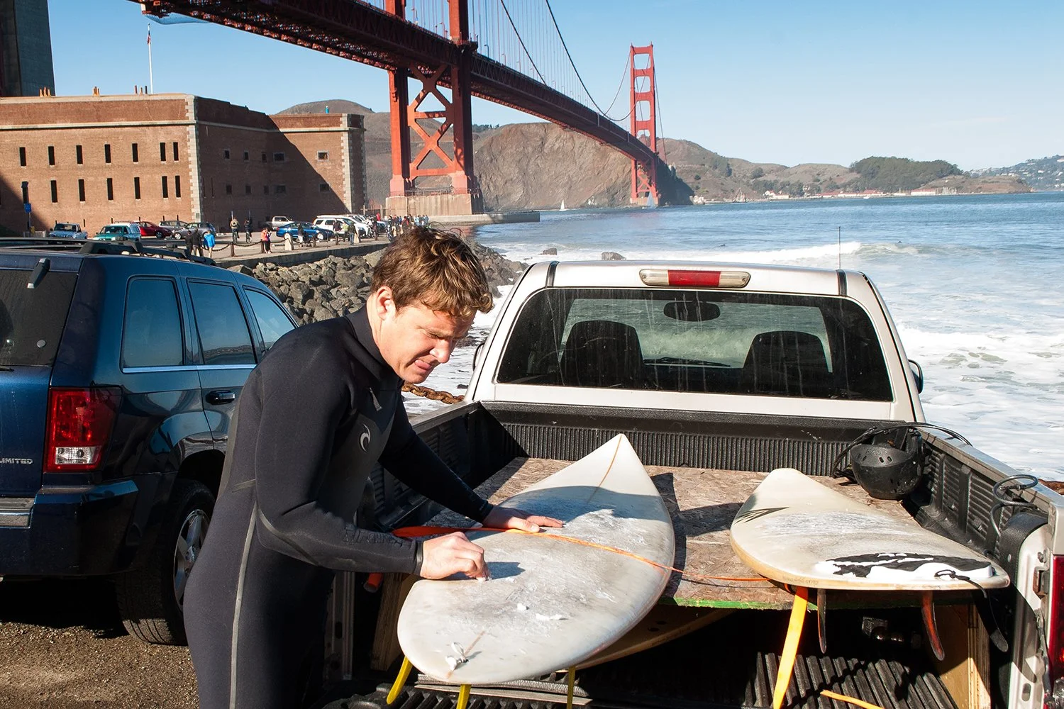 A man wearing a wetsuit preparing a surfboard in the back of a pickup truck on a beach near the Golden Gate Bridge in San Francisco, California.