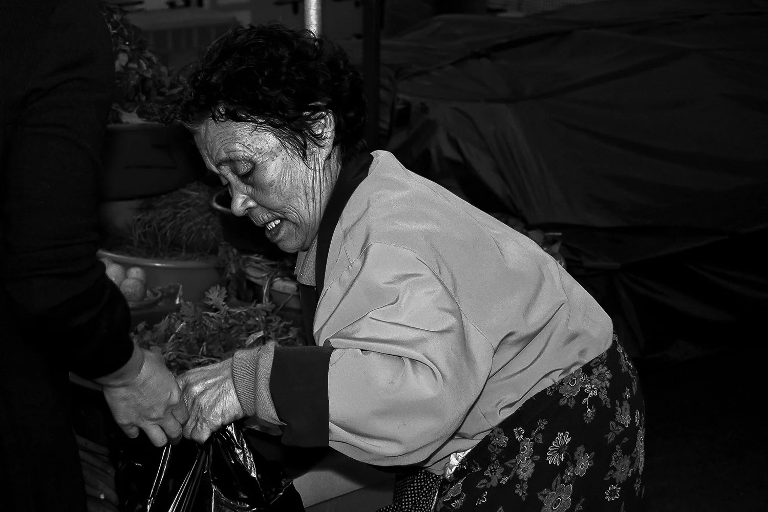 An elderly woman with curly hair is smiling while shopping, holding a bag of produce.