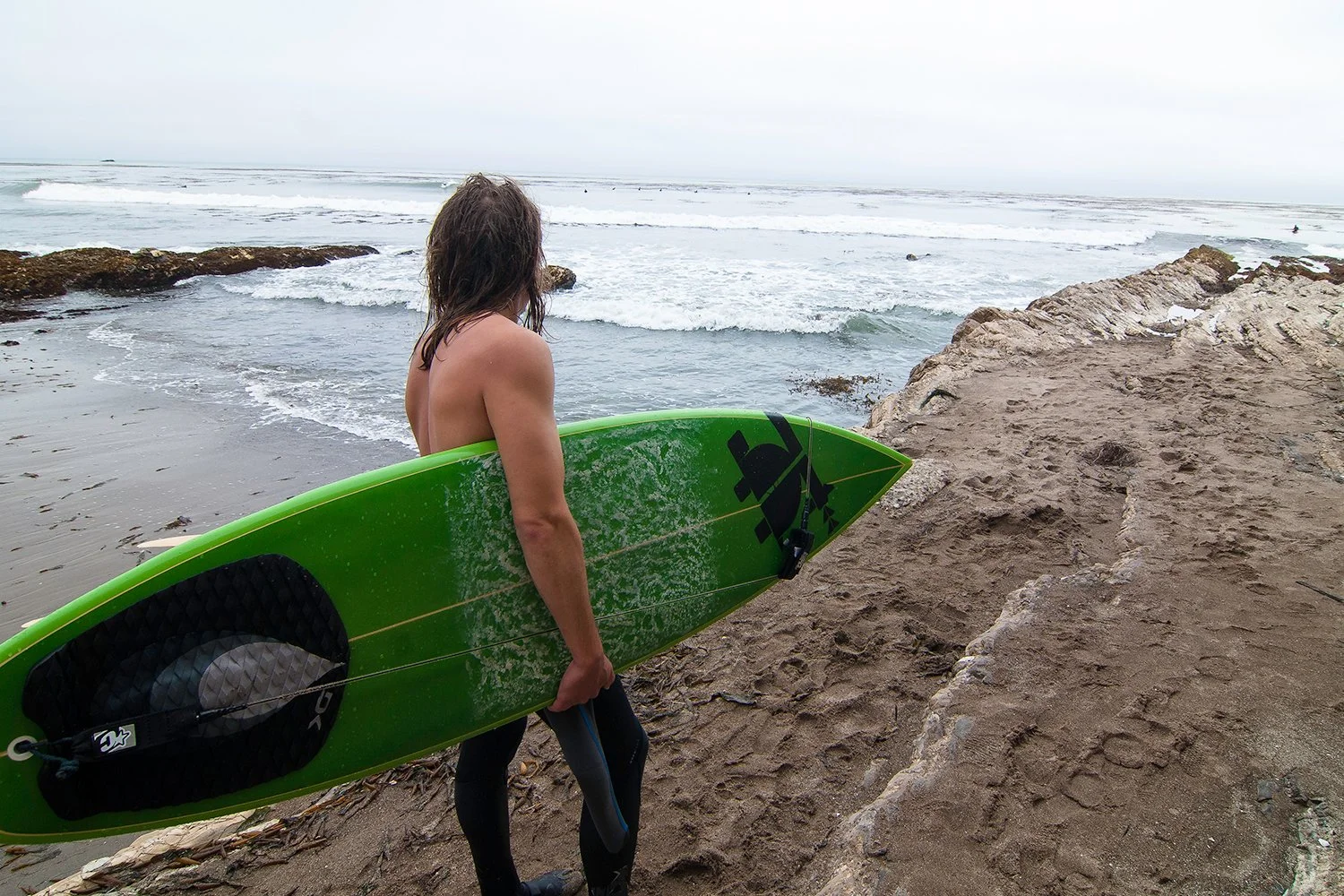 Person with long hair, shirtless, holding a green surfboard, walking towards the ocean on a rocky beach.