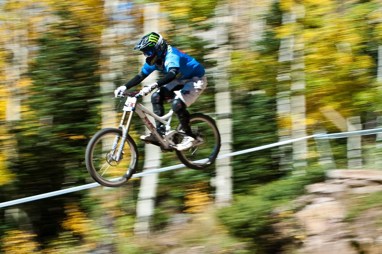 A mountain biker wearing a helmet and blue jersey riding on a trail through a forest with blurred autumn leaves.