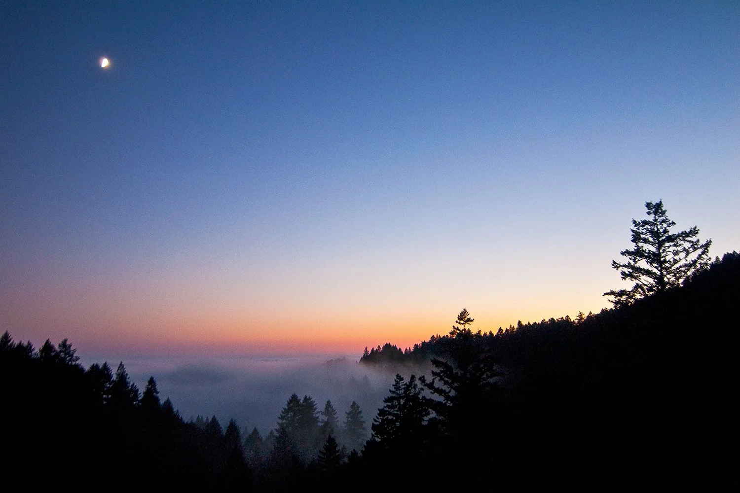 A scenic view of a mountain landscape at sunset, with a crescent moon in the sky and silhouettes of trees in the foreground, with a gradient of colors from purple to orange.