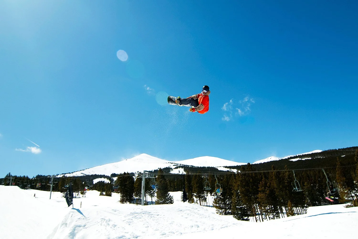 A person snowboarding in mid-air on a snowy mountain, wearing a red jacket, with snow-covered peaks and a clear blue sky in the background.