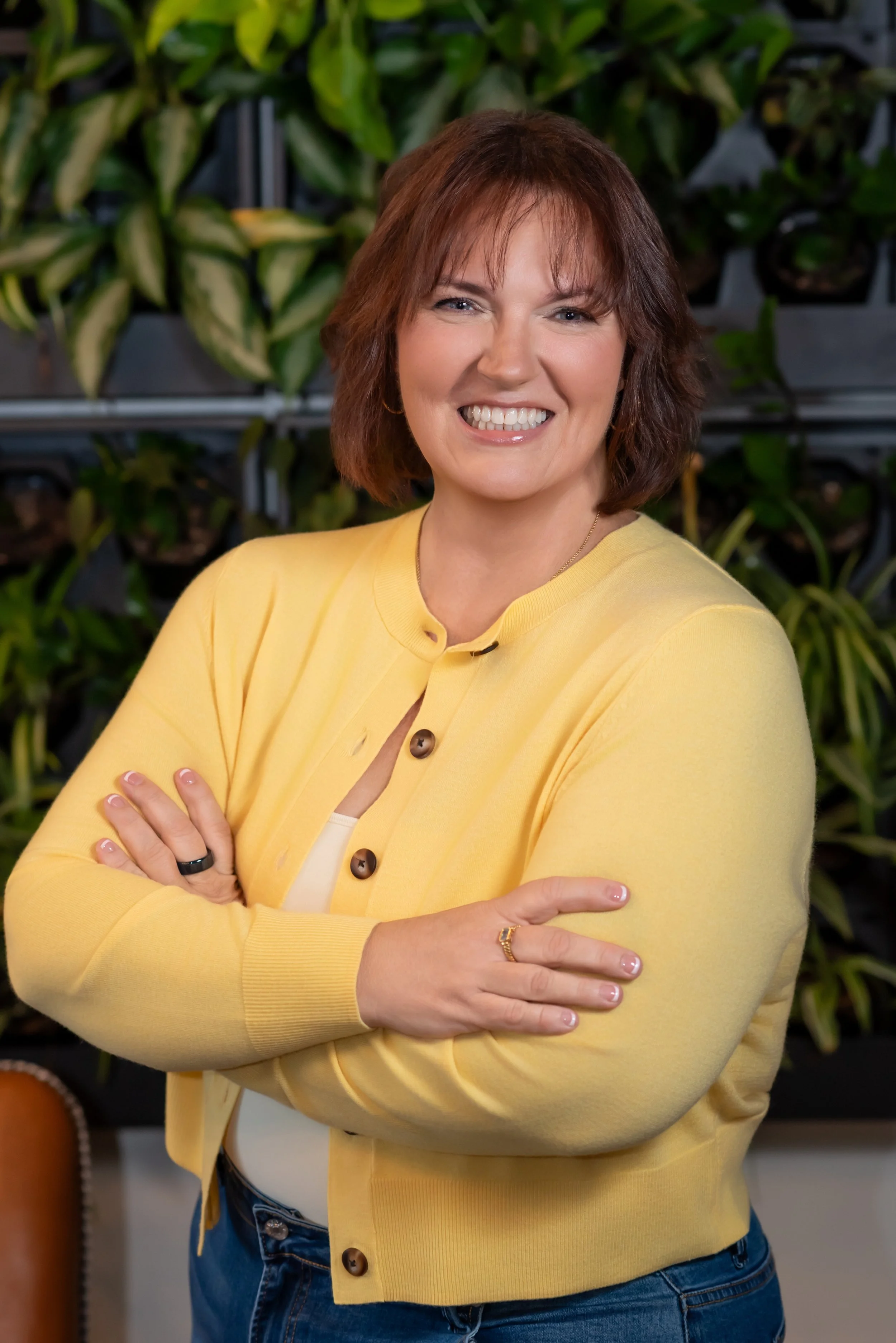 A woman with short brown hair, wearing a yellow cardigan and blue jeans, smiling with arms crossed, standing in front of green plants.